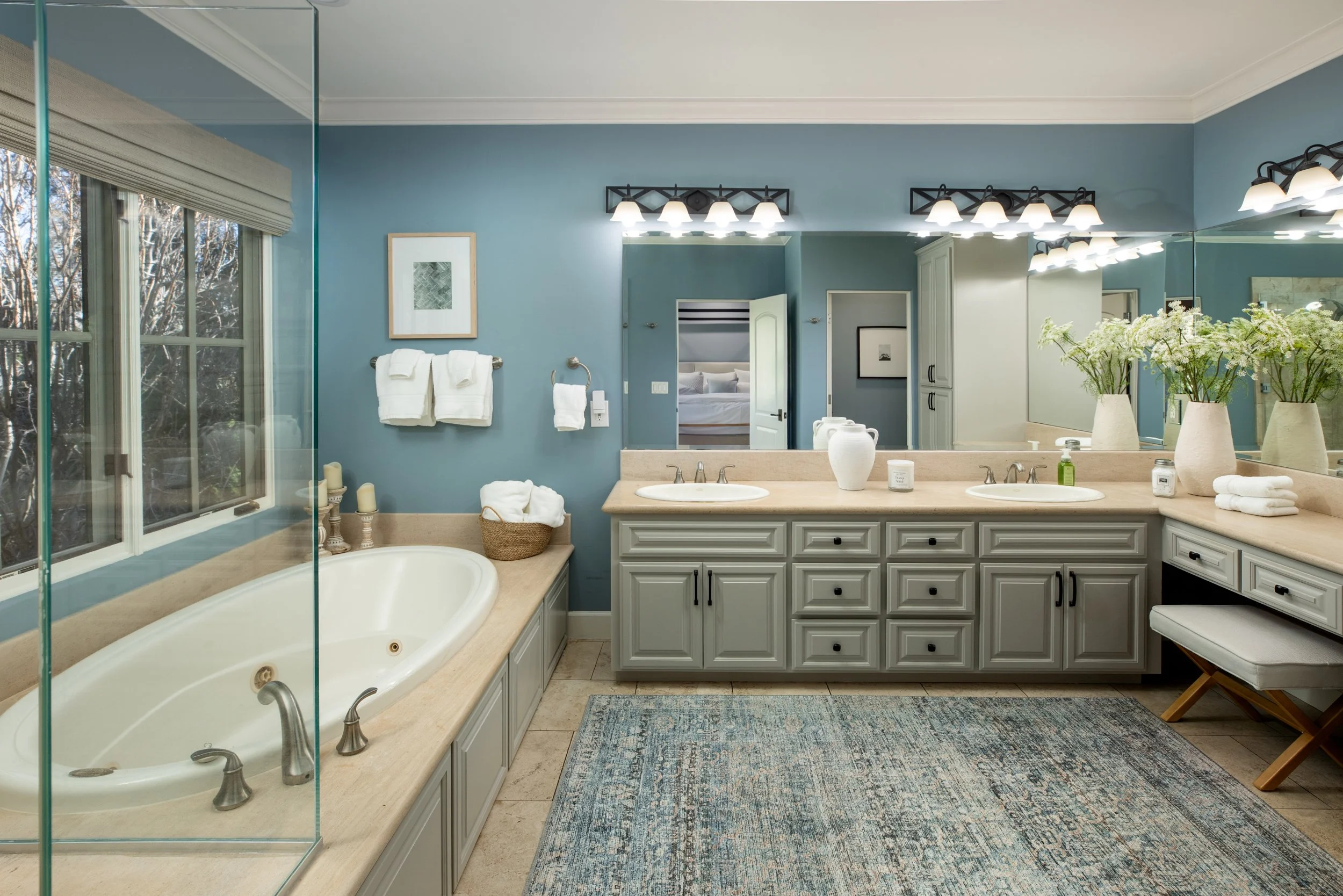 A spacious bathroom featuring a large bathtub, a double sink vanity with a mirror, a window with outdoor trees, and decorative towels and vases with flowers.