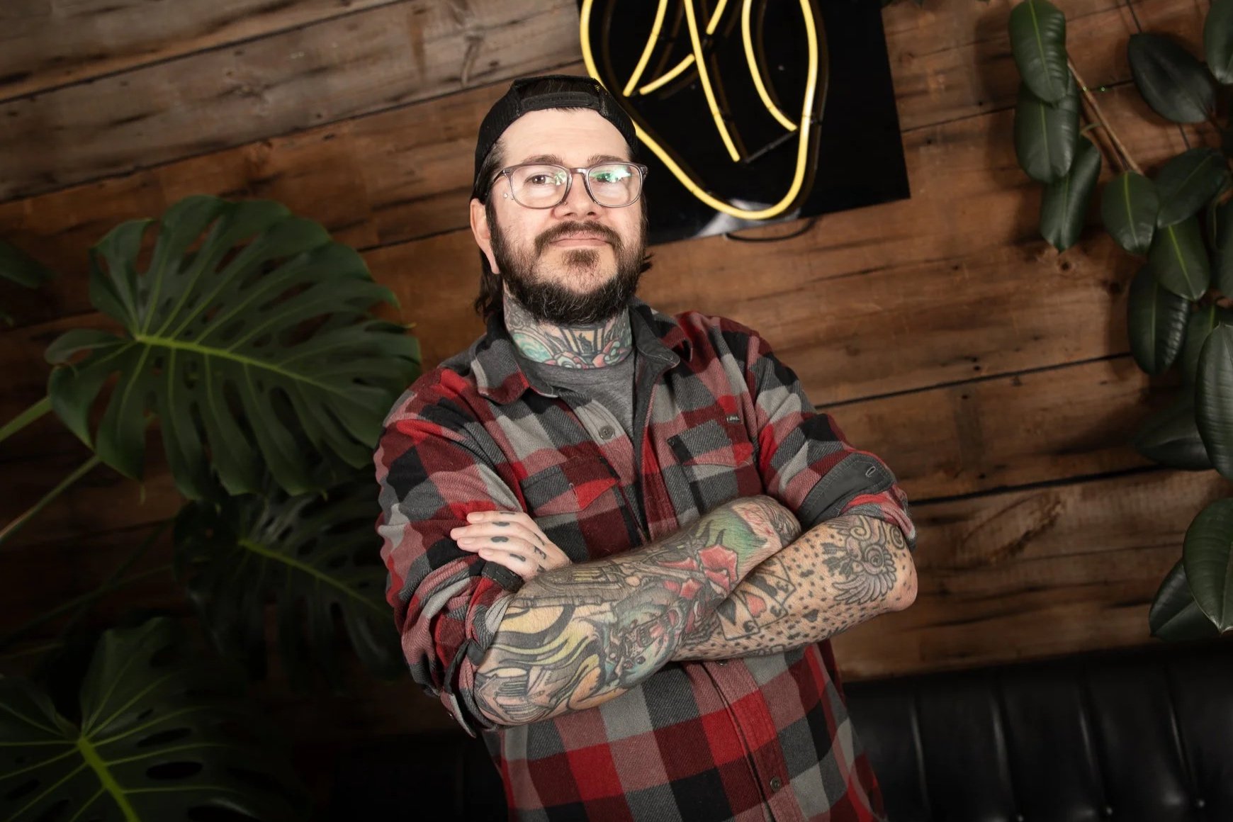 A man with a beard, glasses, and tattoos wearing a black baseball cap and a red plaid shirt, standing with arms crossed in front of a wooden wall and tropical plants, with a neon light in the background.