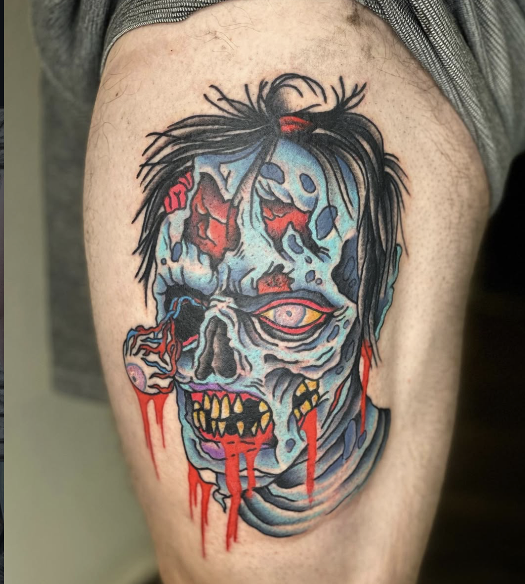 A colorful tattoo of a zombie-like face with multiple eyes, sharp teeth, and decaying skin, with blood dripping from its mouth and wounds, and messy black hair.