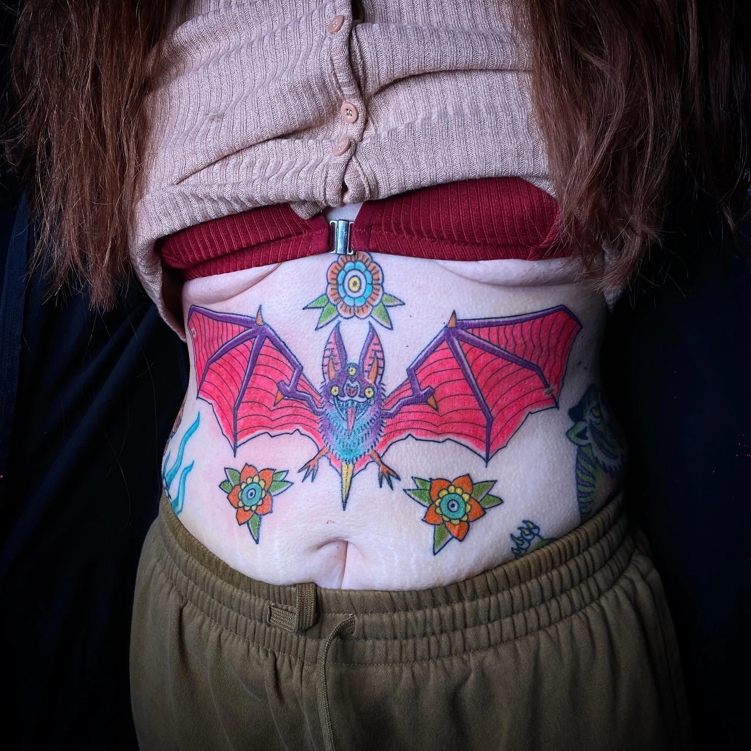 Close-up of a person's midsection wearing a tan ribbed sweater, red pants with a zipper, and gray distressed pants, with long brown hair partially visible.