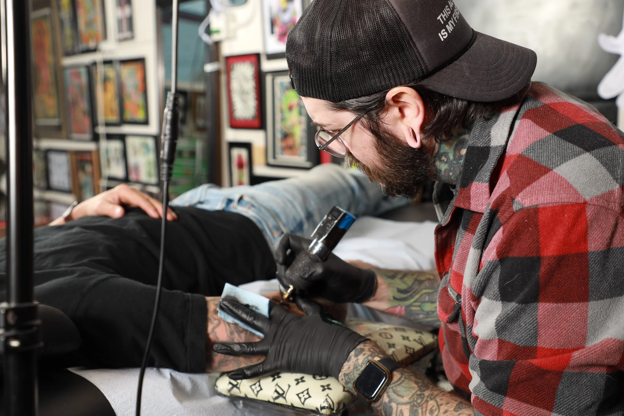 Tattooed person lying on a medical chair receiving tattooing on their arm from a tattoo artist wearing a black cap, glasses, and a red plaid shirt in a tattoo studio with framed art on the walls.