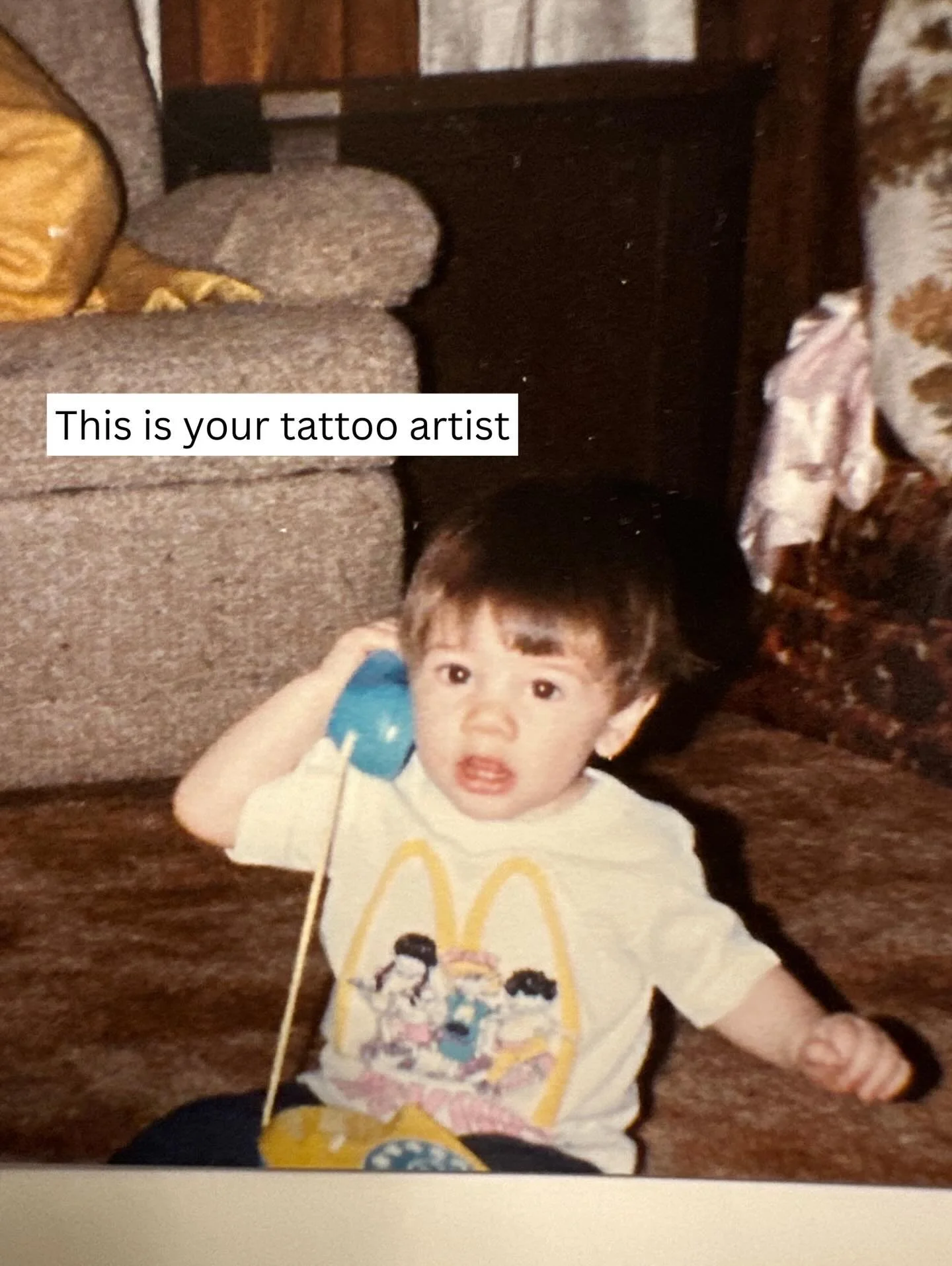 A young boy with dark hair and wearing a McDonald's shirt, holding a blue balloon, sitting in a living room with a brown couch and wooden furniture. He appears to be crying or yawning.