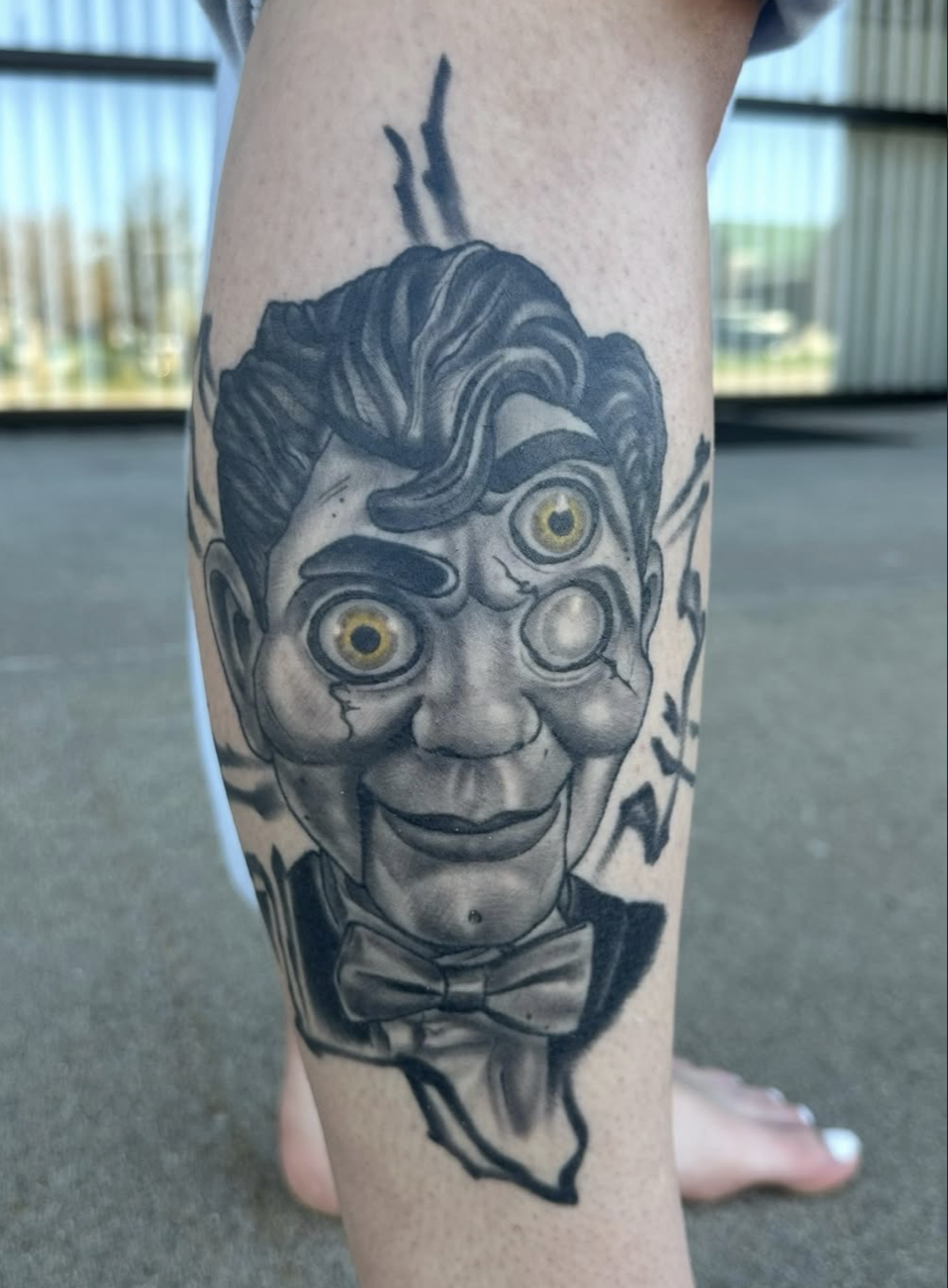 Tattoo of a clown with exaggerated facial features, including large yellow eyes, a big smile, and a bow tie on a person's leg.