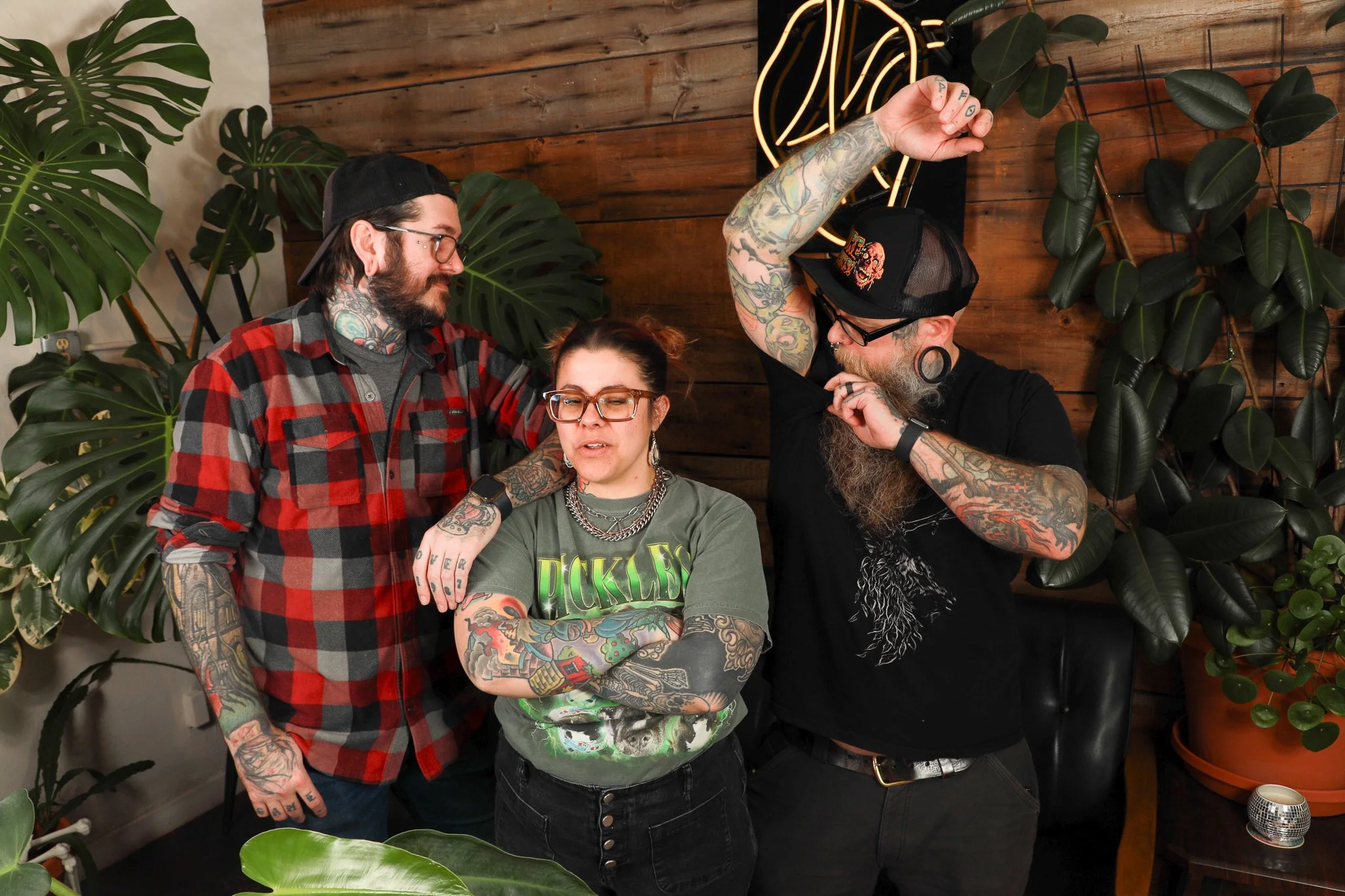 Four people stand together in front of a wooden wall with leafy green plants. The group includes two men with tattoos and glasses, one woman with glasses and tattooed arms, and another man with tattoos, a nose ring, and a hat, gesturing with his arm raised. They are all casually dressed.