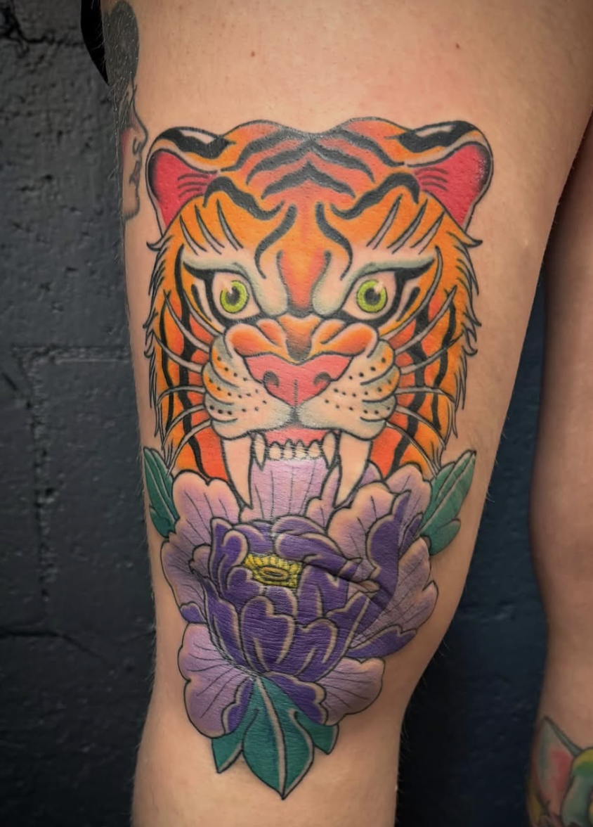 Tattoo of a fierce tiger's face with green eyes and orange, black, and white fur, positioned above a purple flower with green leaves.
