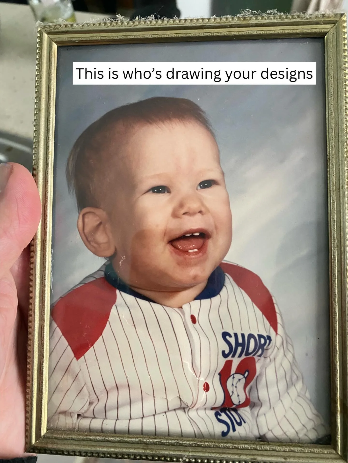 A framed photo of a smiling baby with short hair, wearing a baseball uniform with red and white stripes, and text on the shirt that says 'SHOP' and the number '16'. The person holding the frame is partially visible with a thumb on the left side of the image.