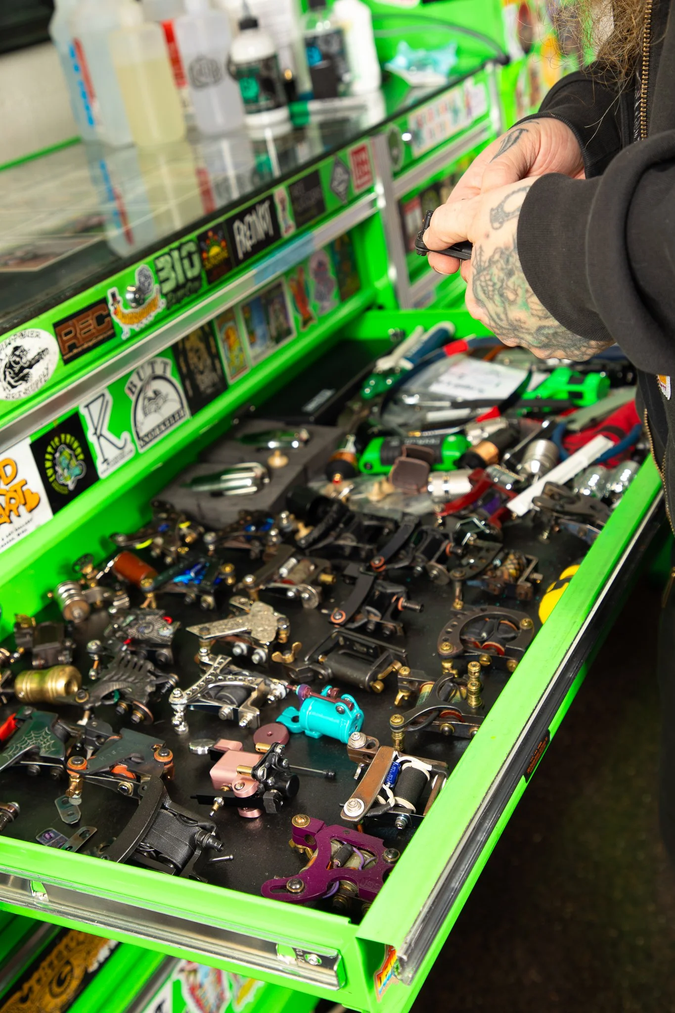 A person with tattoos on their hand is looking through a display case filled with various tattoo machine parts.