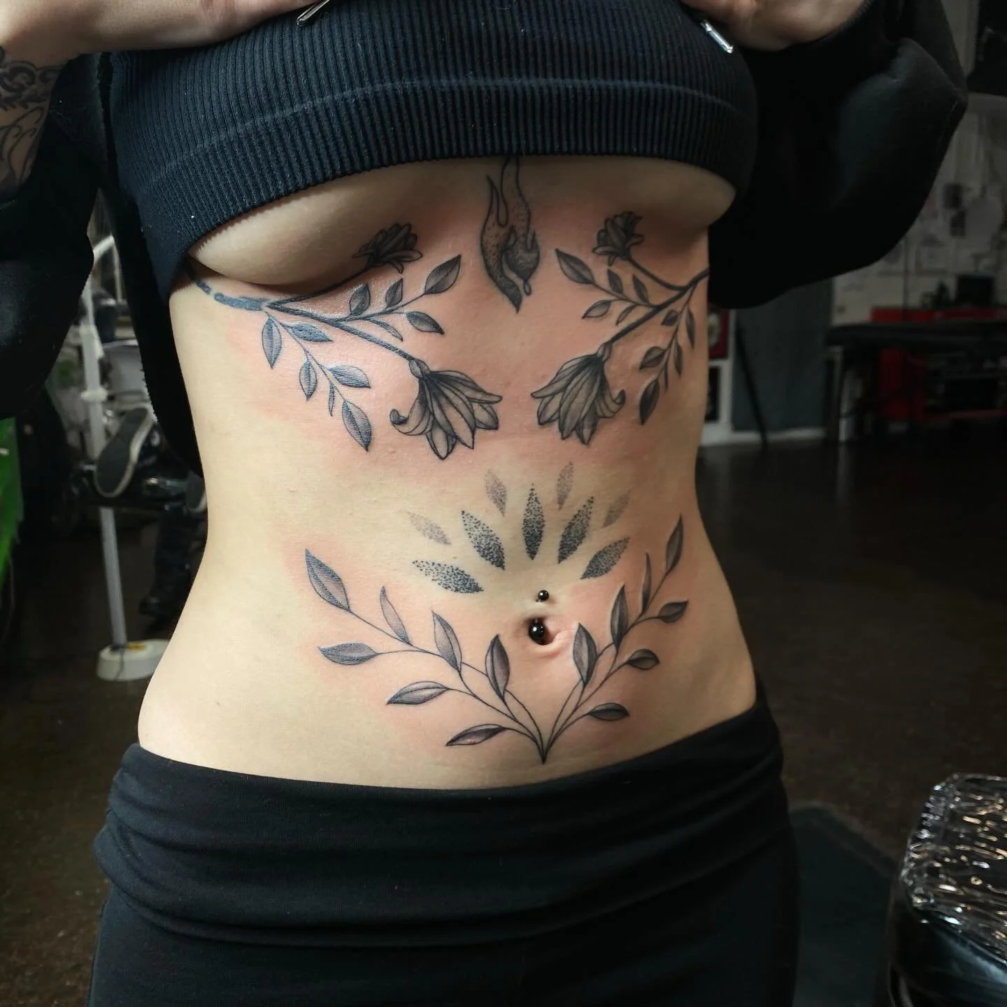 A person's torso with fresh black and gray floral tattoos on the lower chest and abdomen, including leafy branches and hanging flowers, along with a belly button piercing.
