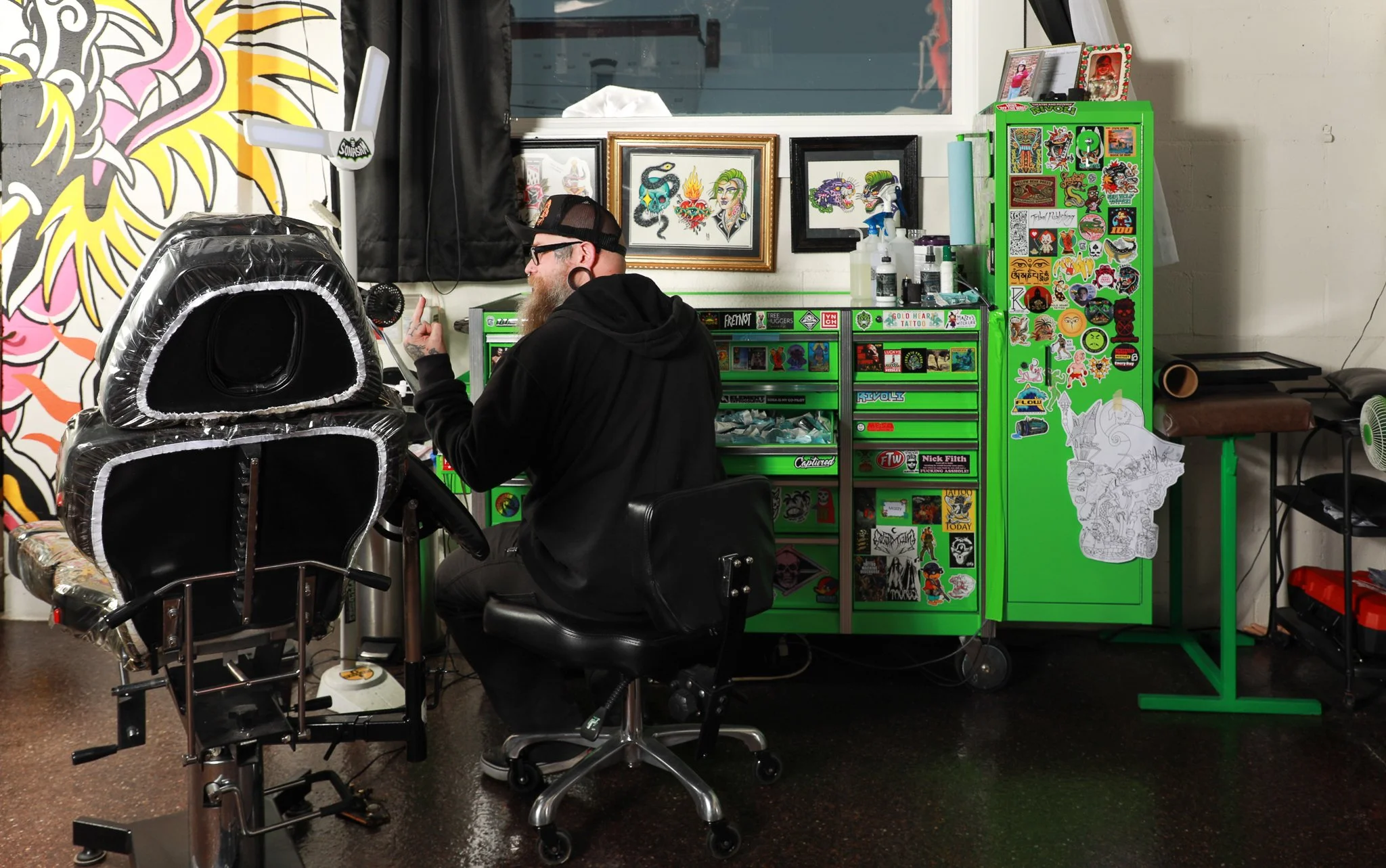 A man with glasses and a cap tattooing a person's leg in a tattoo shop. The shop has colorful artwork, stickers, and tattoo supplies.