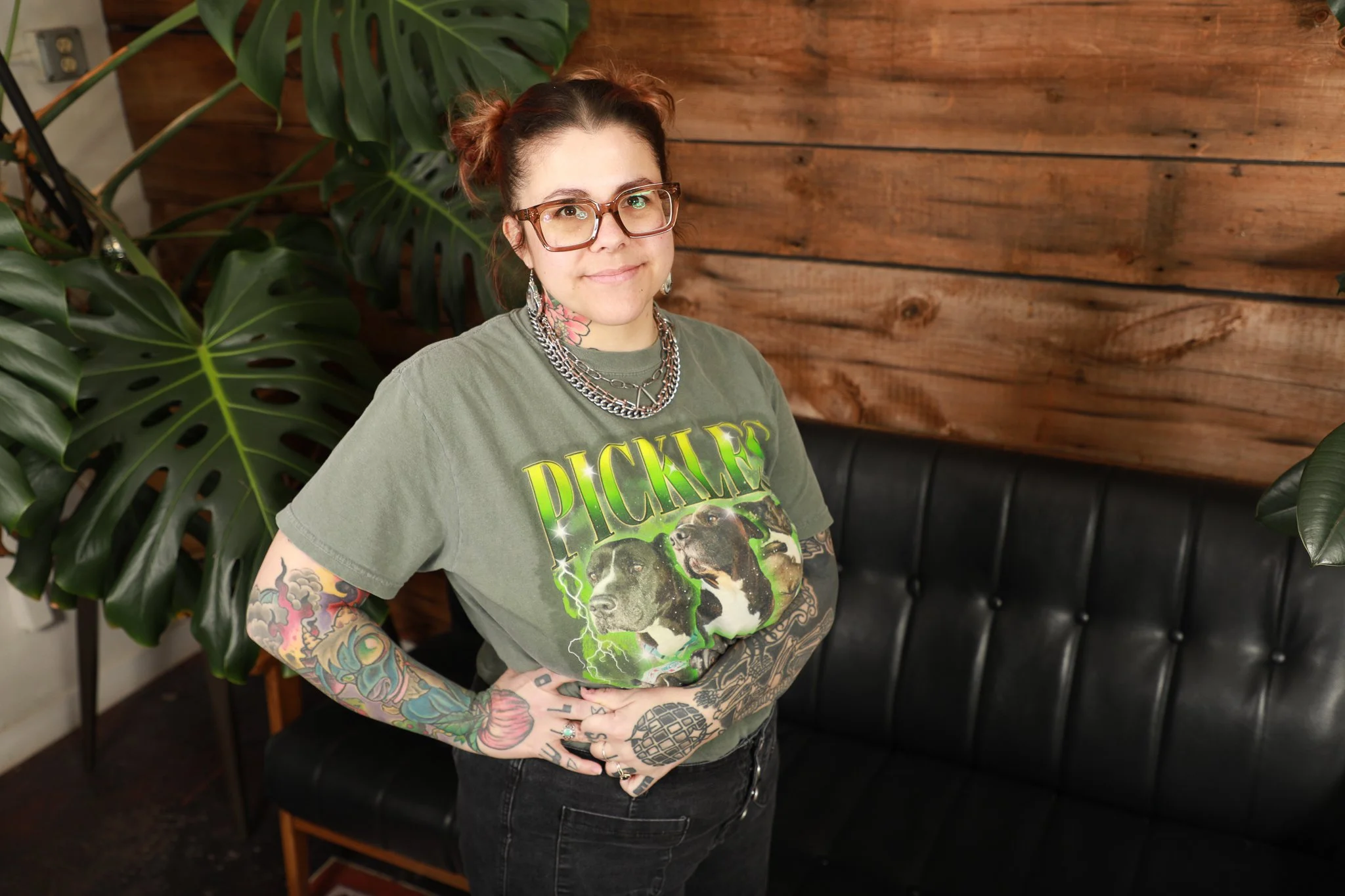 A woman with tattoos, glasses, and a graphic t-shirt standing indoors in front of a wooden wall and large green plants.