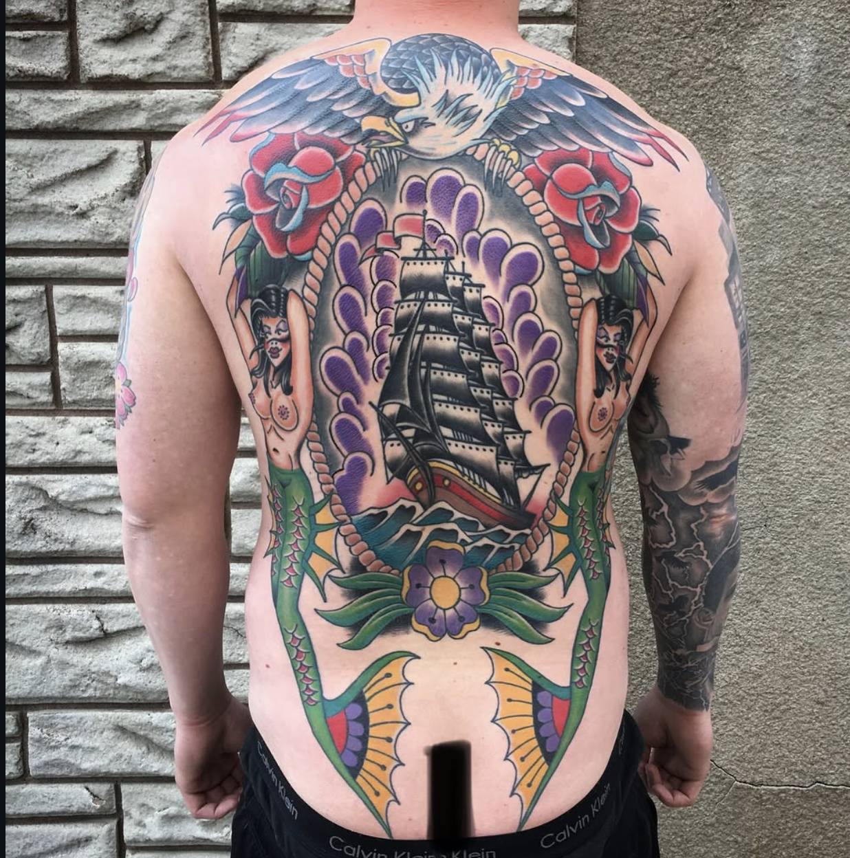 Colorful tattoo on a person's back featuring a ship inside a flower-shaped frame, an eagle at the top, roses and women on the sides, and decorative elements including leaves and other floral designs.