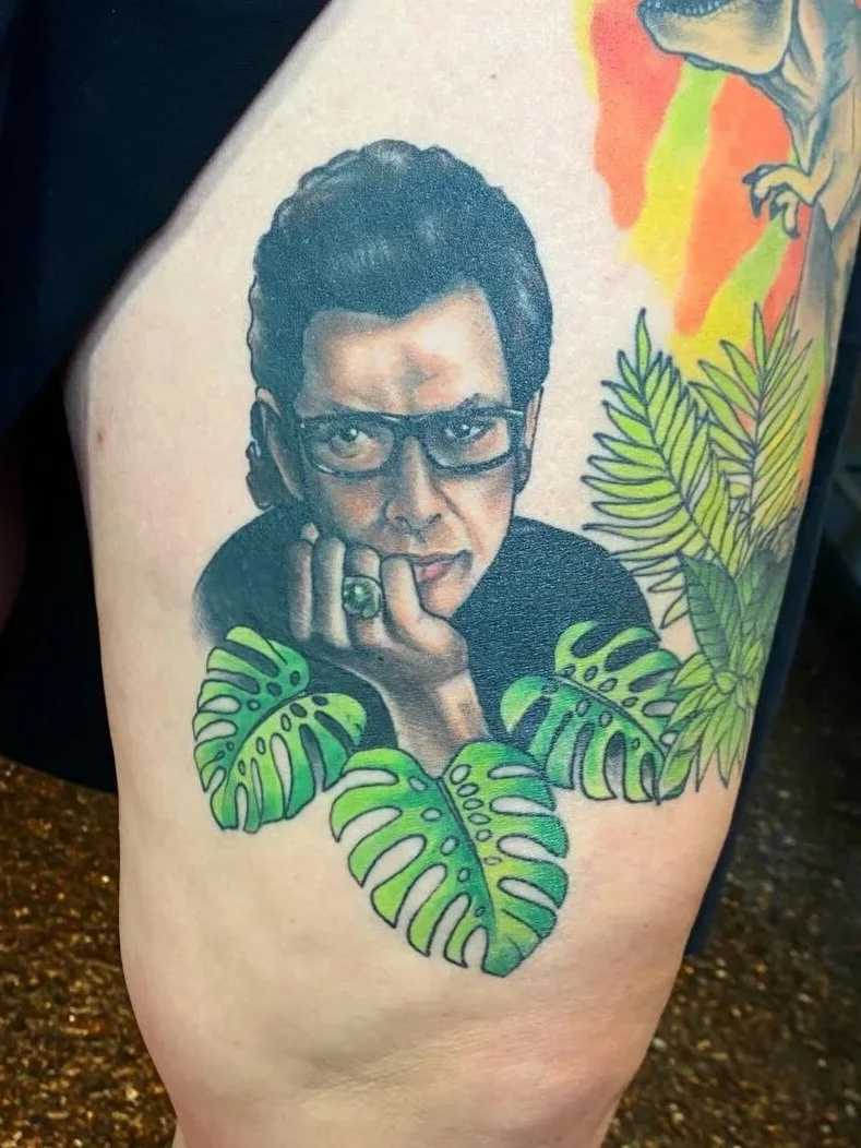 Tattoo of a woman with short dark hair and glasses resting her chin on her hand, surrounded by green tropical leaves.