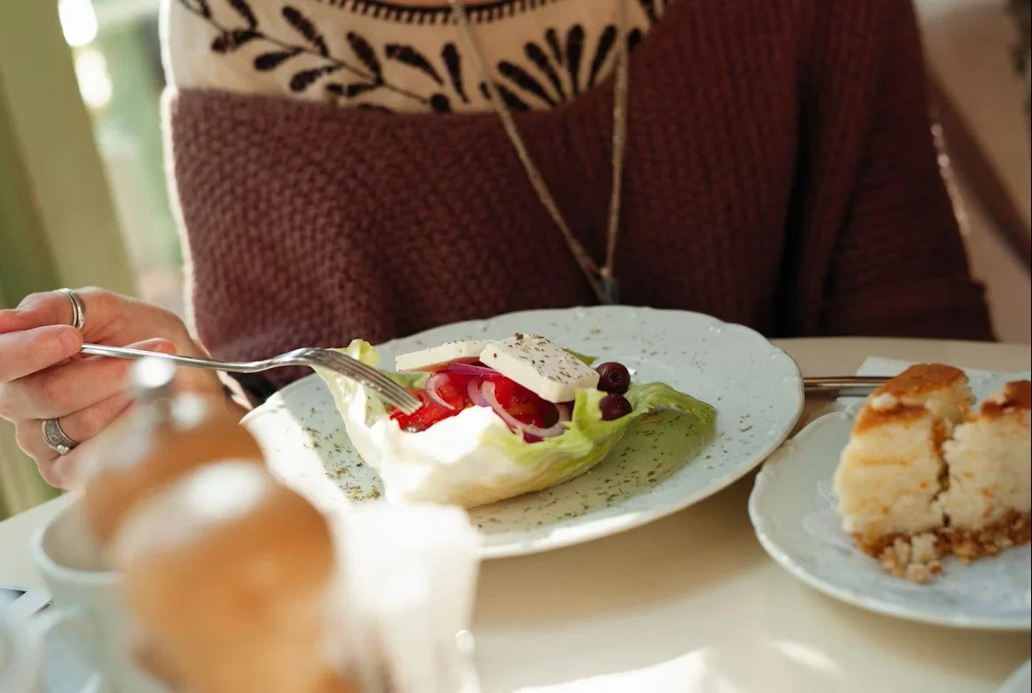 A person eating a Greek salad with feta, tomato, and olives, with a slice of cake on a nearby plate, at a café table.