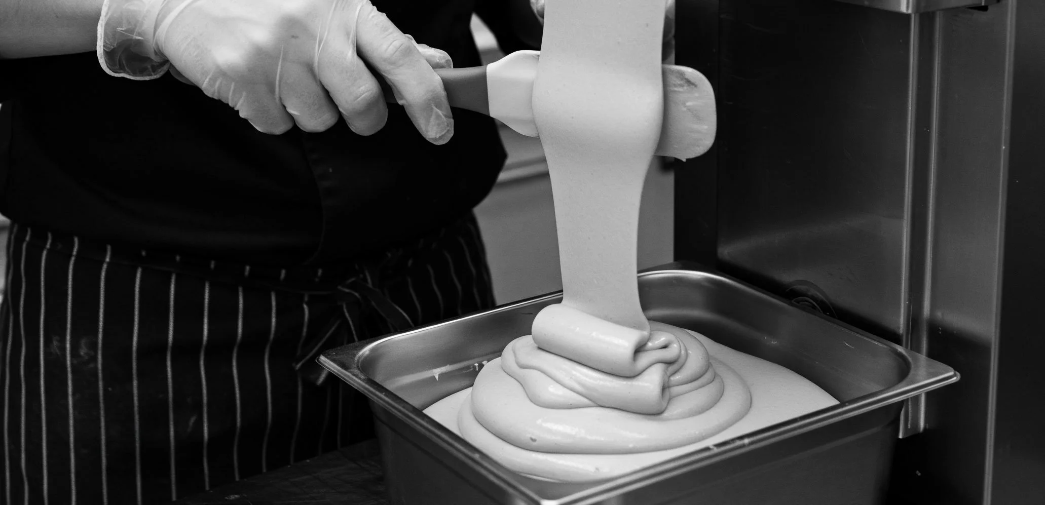 A person wearing gloves is pouring a creamy, smooth batter or mixture into a rectangular baking pan, creating swirls on the surface.