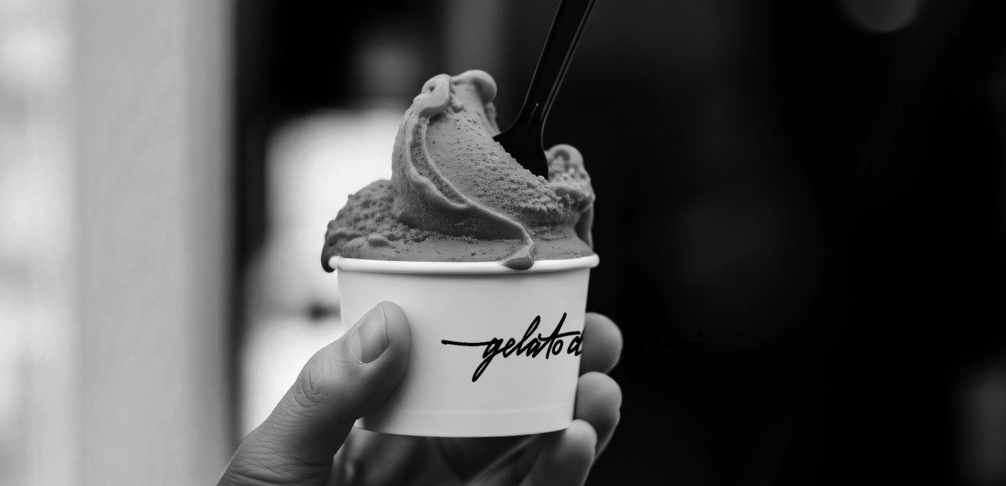 A person holding a paper cup filled with ice cream with a spoon sticking out, written on the cup is 'gelato'.