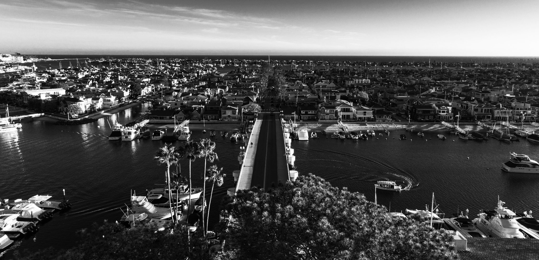 Black and white aerial view of a marina with numerous boats docked, a bridge crossing over water, and residential houses in the background.