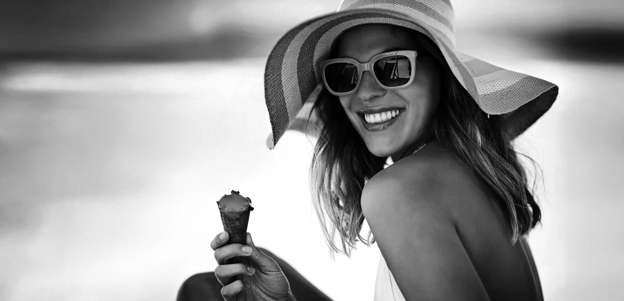 A woman smiling while holding an ice cream cone, wearing sunglasses and a wide-brimmed hat with stripes.