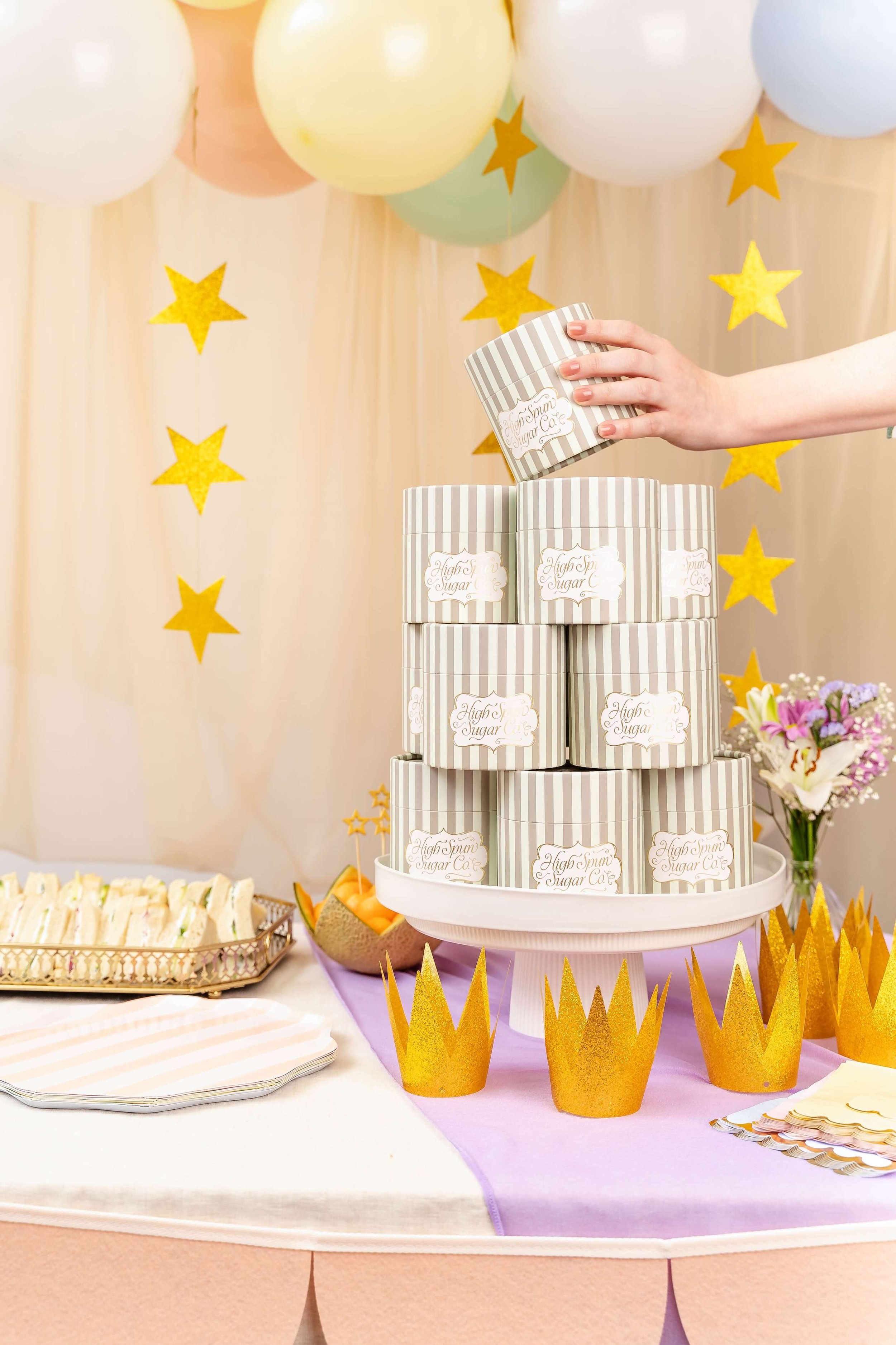 A birthday party table with pastel balloons, yellow star confetti, a tiered cake stand with striped boxes labeled 'High Spun Sugar Co.', gold paper crowns, and a floral arrangement in a basket, all on a table covered with pink and purple decorations.