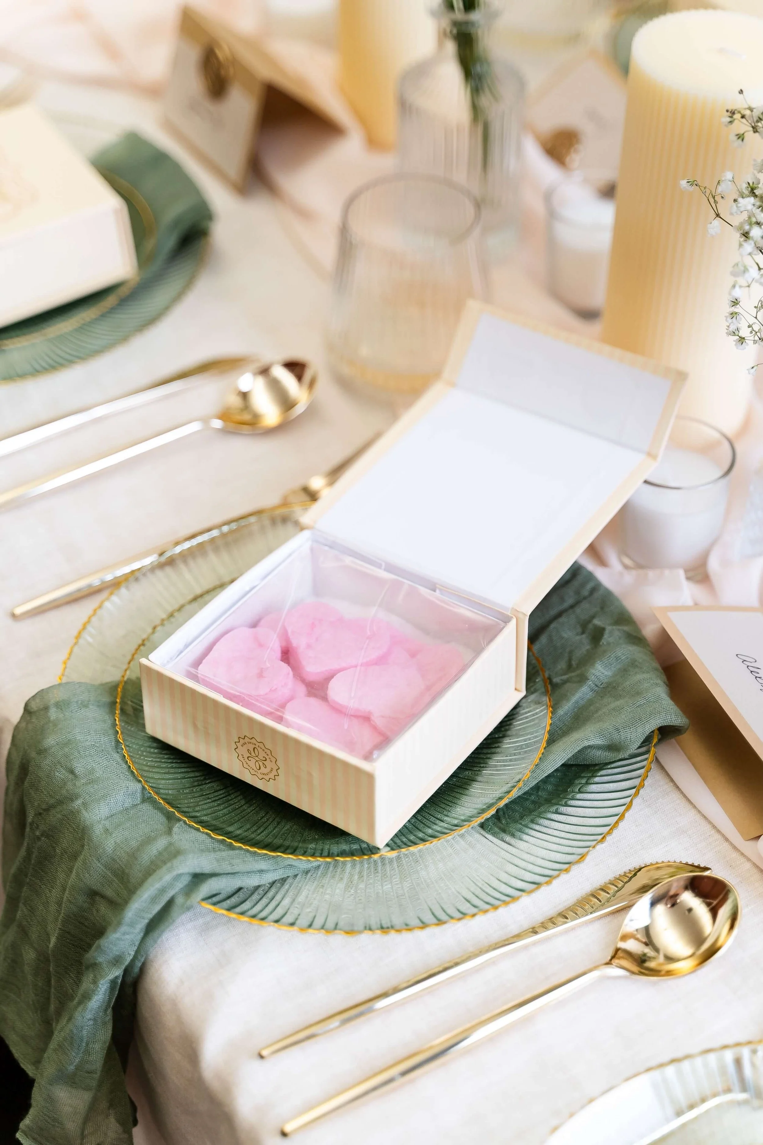 A decorated table with a green charger plate, a white box full of pink heart cotton candy, gold cutlery, and various candles and floral arrangements.