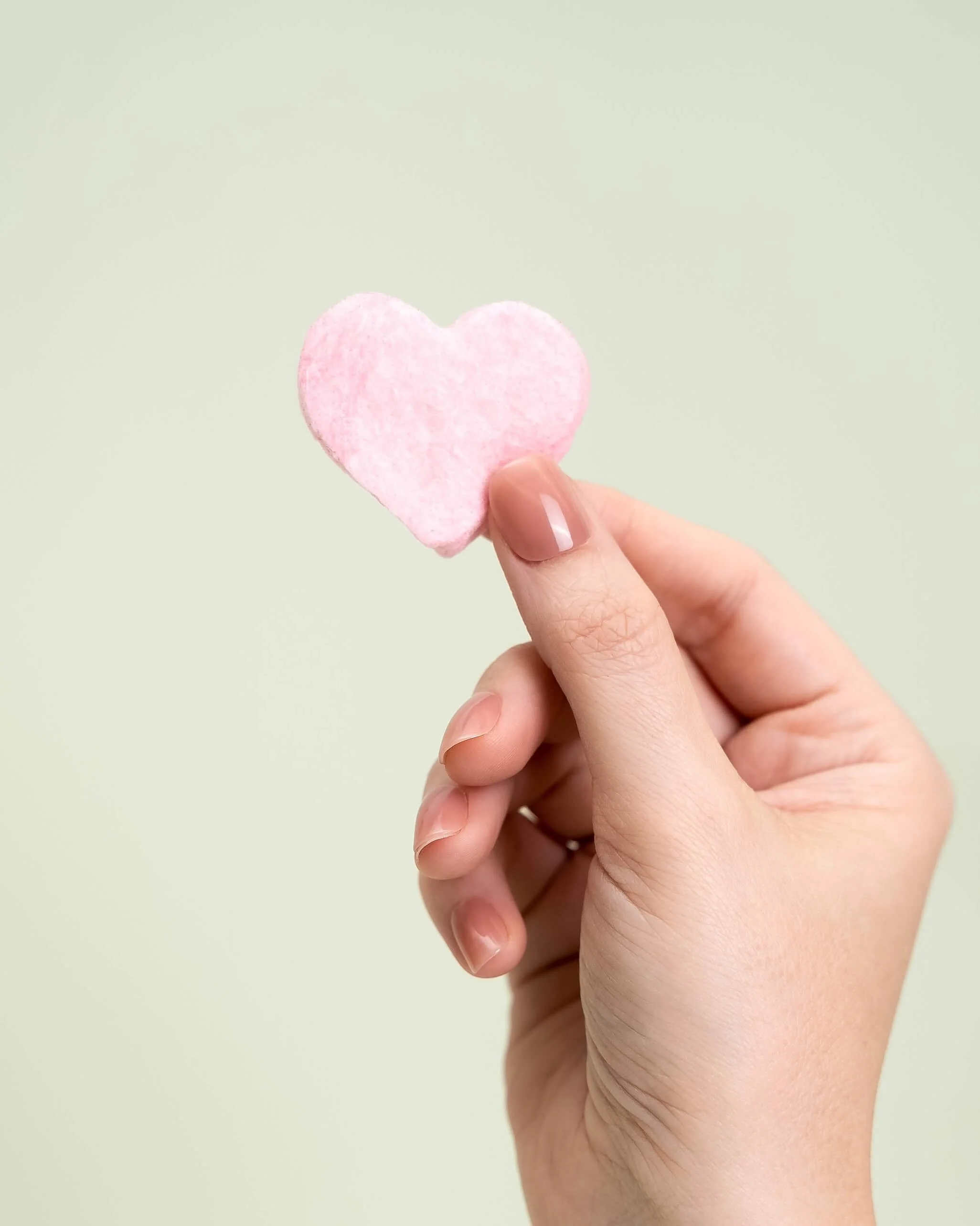 A hand holds a pink cotton candy heart in front of an olive green background