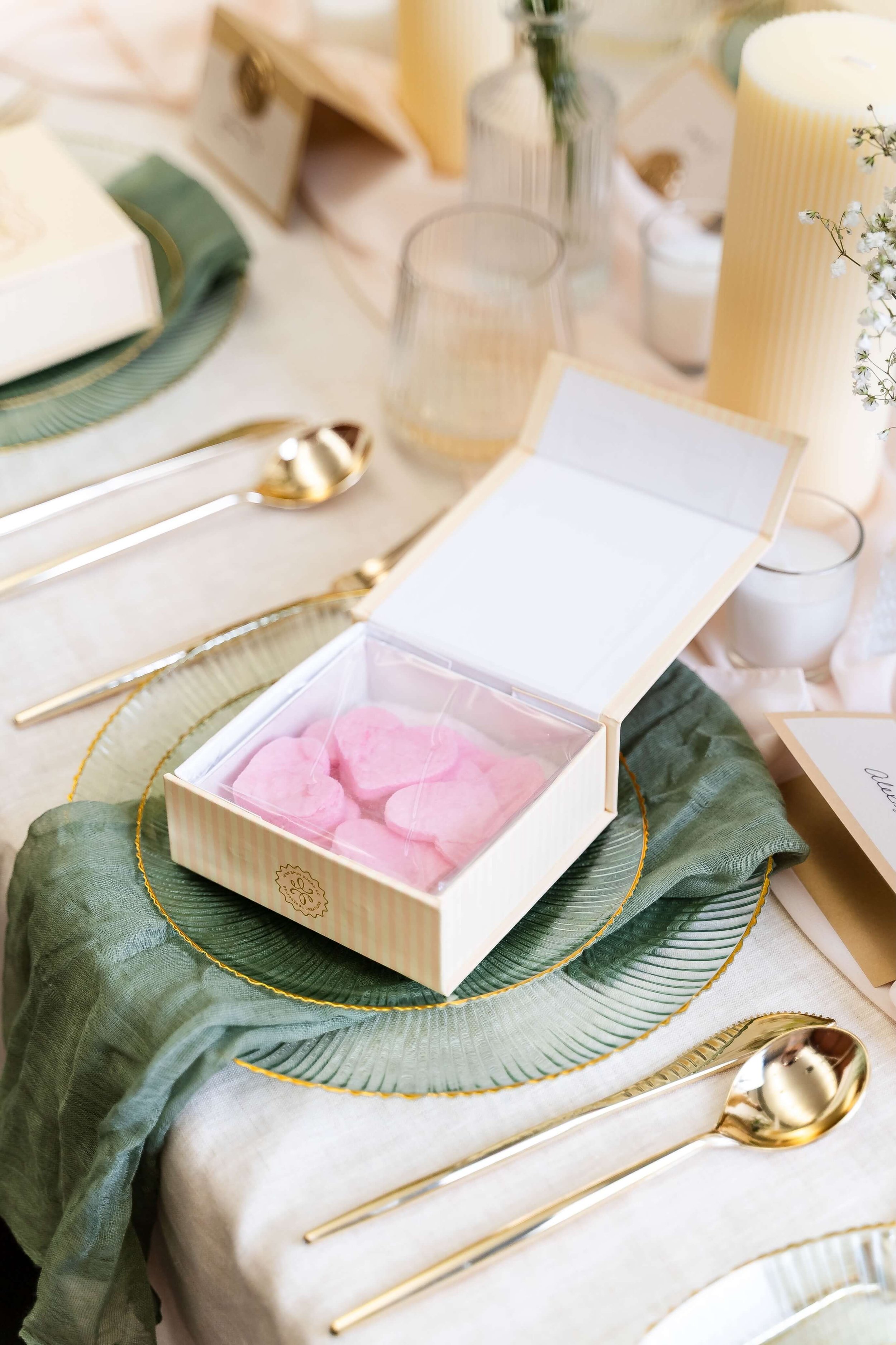 A table set with gold utensils, a green and gold charger plate, a white candlestick, and a box of pink cotton candy hearts, decorated with a green cloth and surrounded by other candles and decorative items.