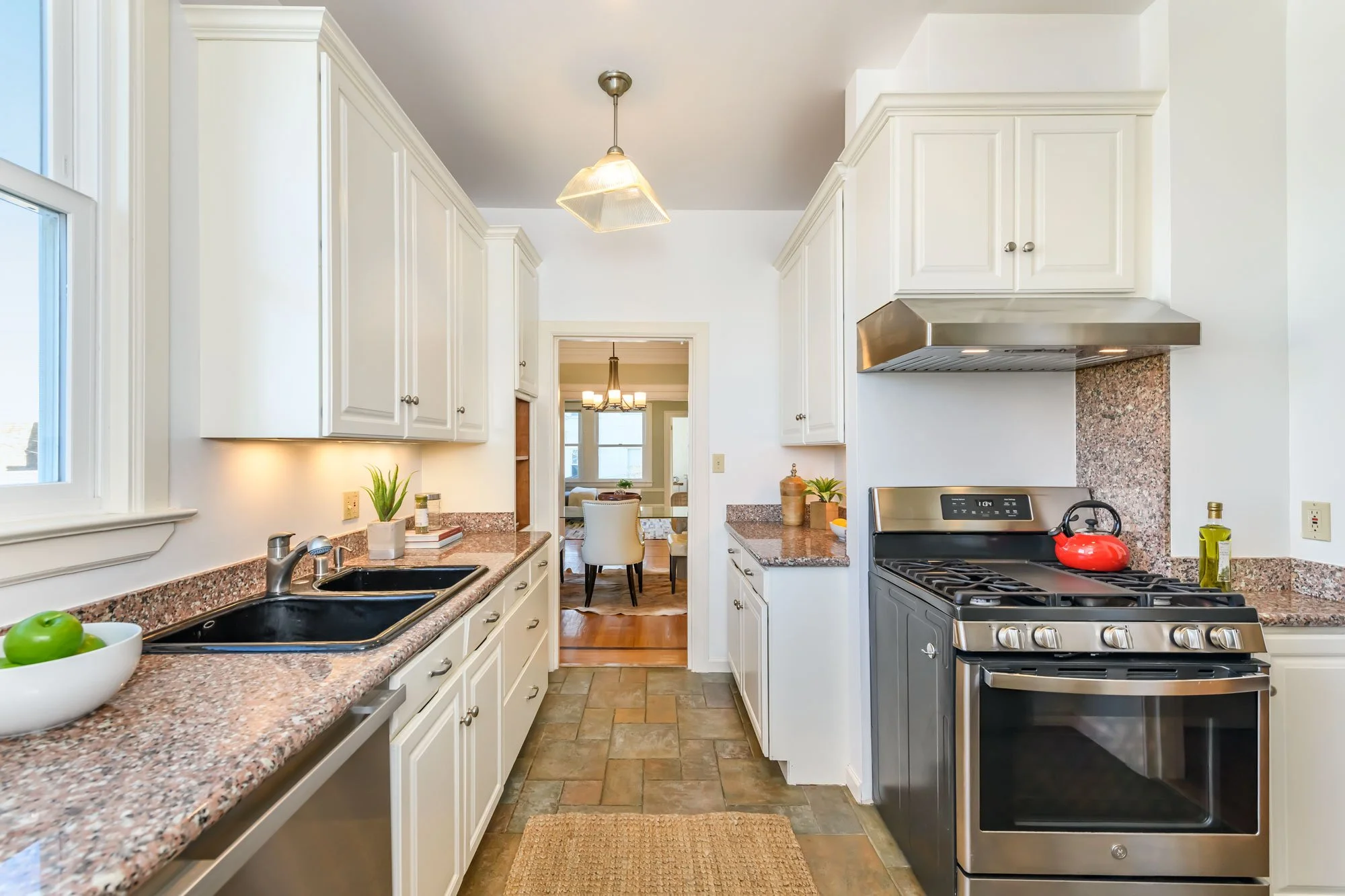 A kitchen with white cabinets, granite countertops, a double sink, a stove with a red kettle on top, and a view into a dining area with chairs and a table.