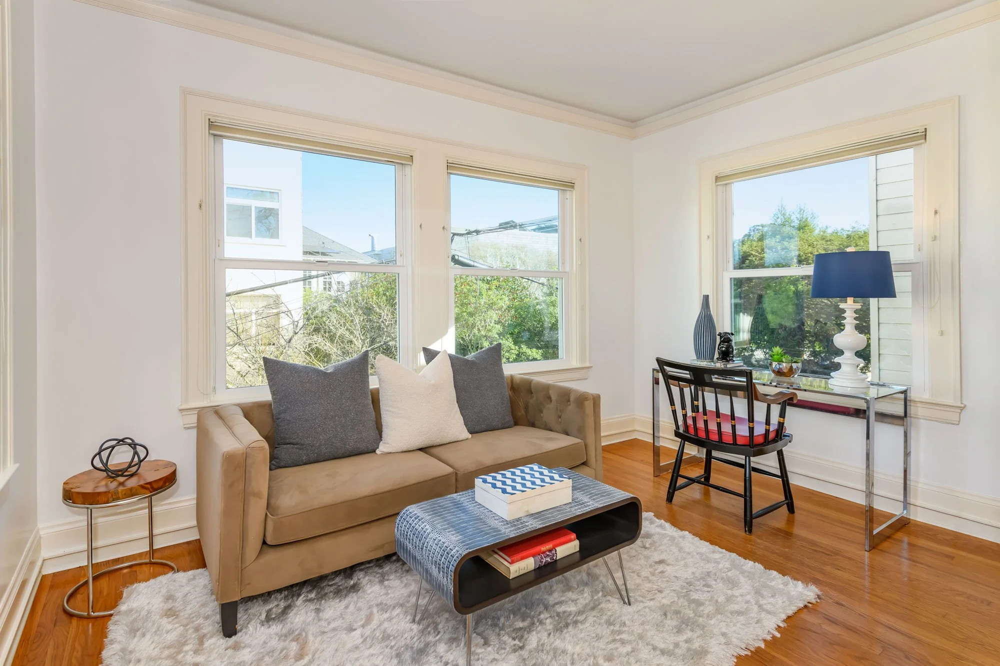 Sunny living room with beige sofa, gray and white pillows, glass coffee table, white shag rug, black side table, and black desk with a blue lamp and decorative items, three large windows showing trees and neighboring houses.