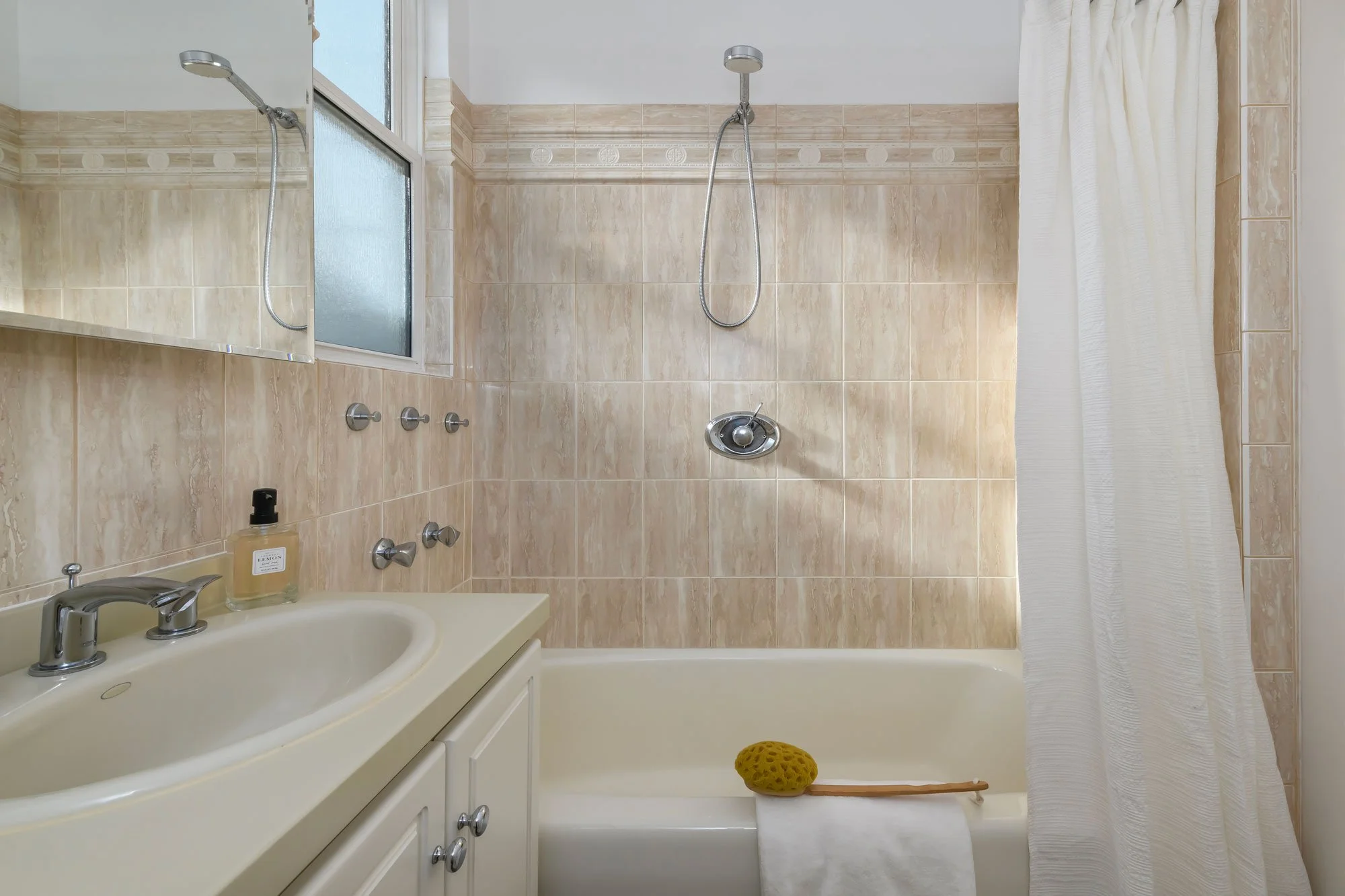 Bathroom with beige tile walls, a white bathtub, and a shower head. There is a white sink with a silver faucet, a mirror, and a bottle of hand soap on the counter. A white towel and a yellow bath sponge are on the bathtub, with a shower curtain parti