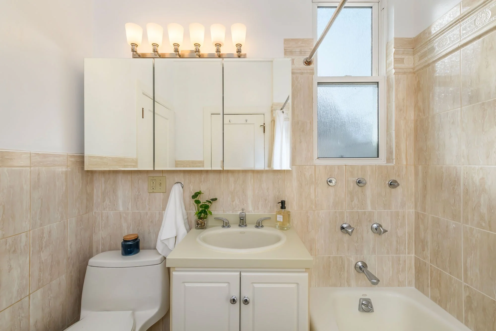 Small bathroom with beige tiled walls, a white toilet, a white sink vanity with a plant and soap dispenser, a mirrored cabinet, a frosted glass window, and a bathtub with wall-mounted fixtures.