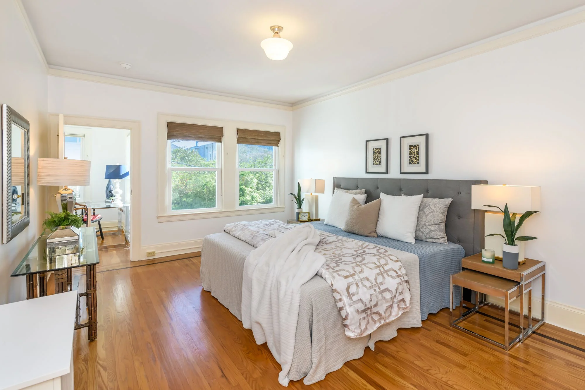 A bedroom with a large bed, gray headboard, white and beige pillows, white blankets, wooden nightstand with a lamp and plant, hardwood floor, and three windows with blinds letting in natural light.