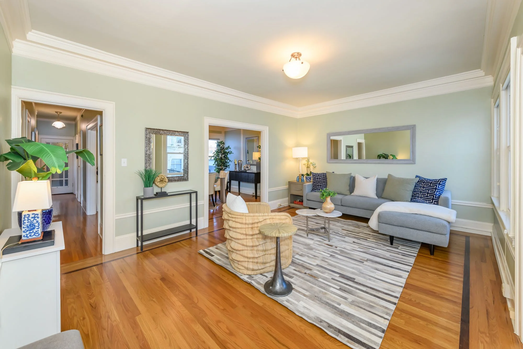 Living room with a gray couch, striped area rug, beige armchair, and wooden side table with lamp; light green walls, white moldings, large mirror, and windows with curtains.