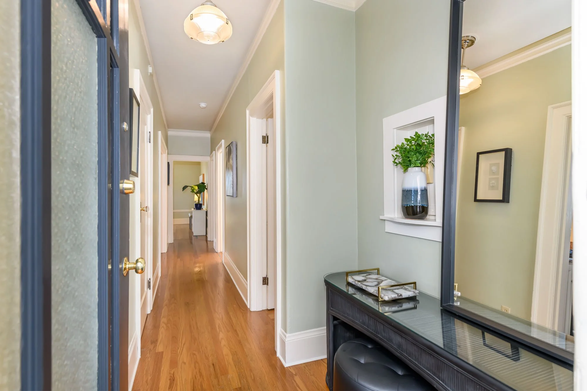 A hallway with green walls and white trim, hardwood floors, a large mirror on the right, a black console table with decorative trays, framed art and a potted plant, and ceiling lights.