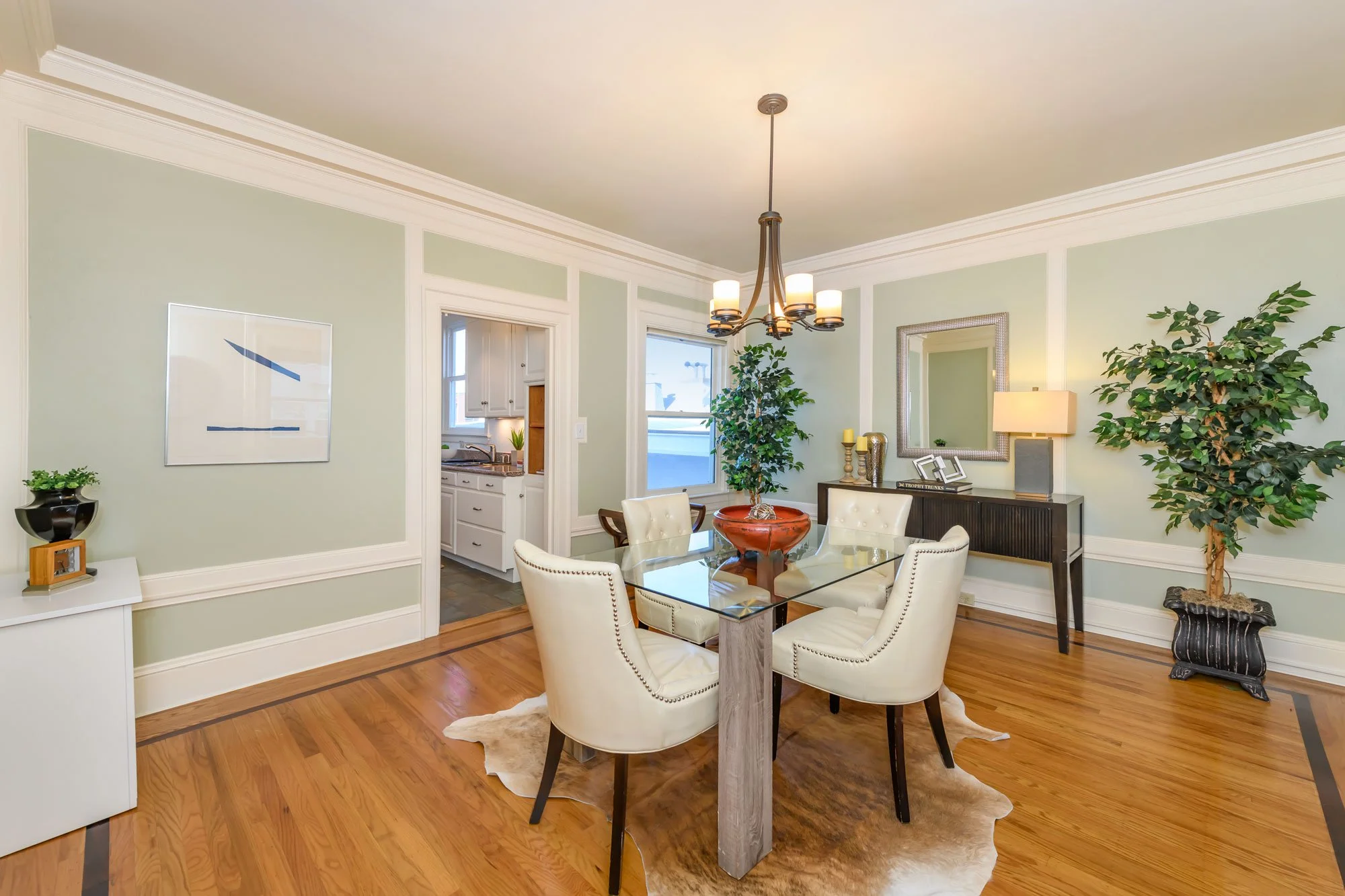 Dining room with beige chairs around a glass table, a chandelier, potted plants, and wall art, with a view into a kitchen.