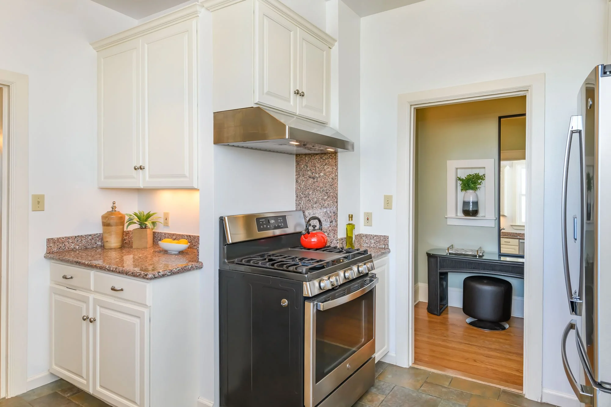 Kitchen with white cabinets, black stove with red kettle, granite countertops, and view into a small room with a black vanity, black stool, and a potted plant on a window sill.