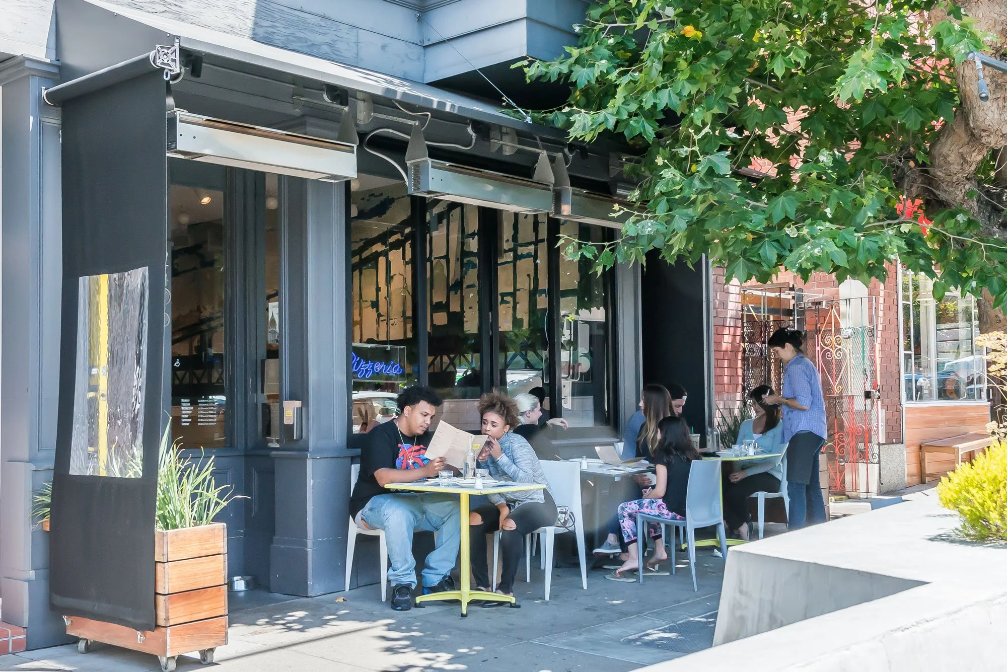 People dining at an outdoor restaurant on a city sidewalk, with a tree providing shade and a brick building in the background.