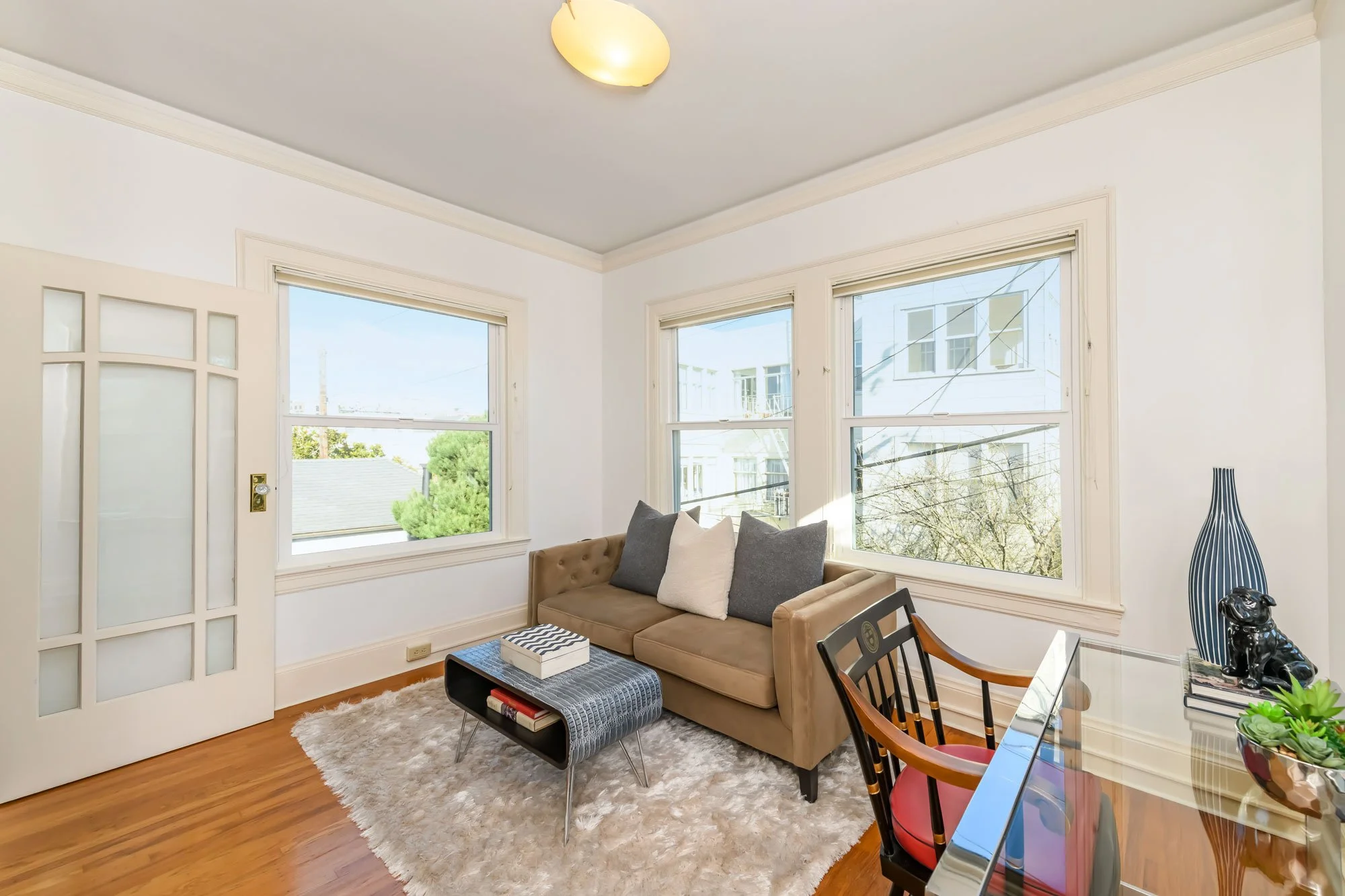 Living room with white walls, hardwood floor, a beige sofa with three decorative pillows, a black coffee table, a white shag rug, a desk with a glass top, and large windows letting in natural light.