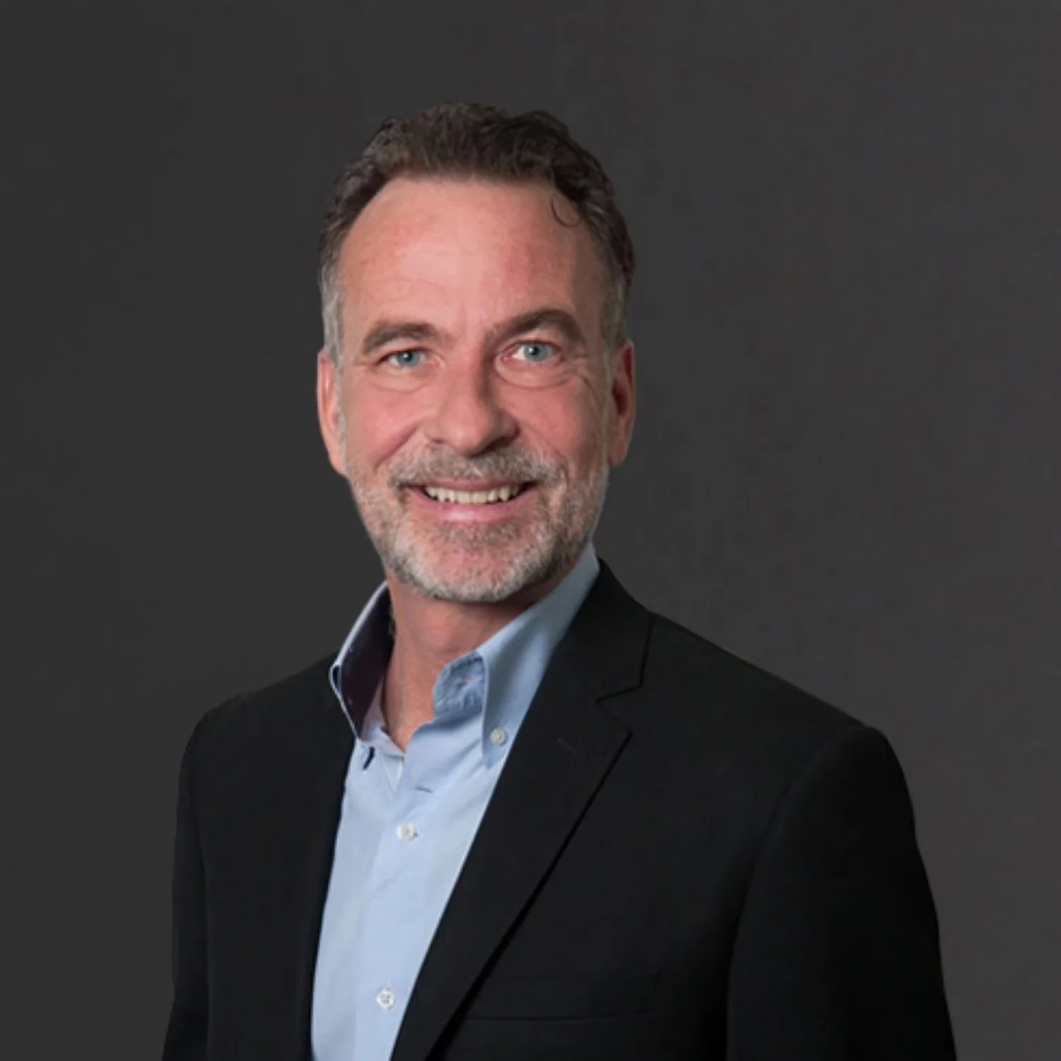 A middle-aged man with salt-and-pepper hair and a beard is smiling while looking at the camera. He is wearing a black blazer over a light blue dress shirt, and there is a dark gray background.