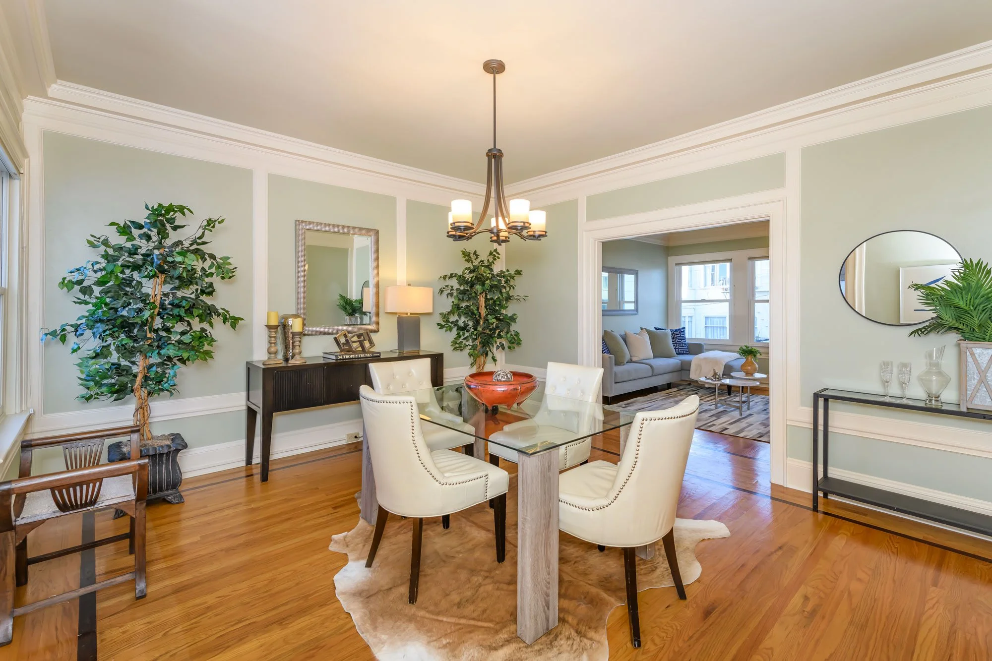Bright dining room with a glass table, four white upholstered chairs, and a cowhide rug. Decor includes a chandelier, mirror, plants, and side tables with lamps. Adjacent living room with large windows and sofa visible in the background.