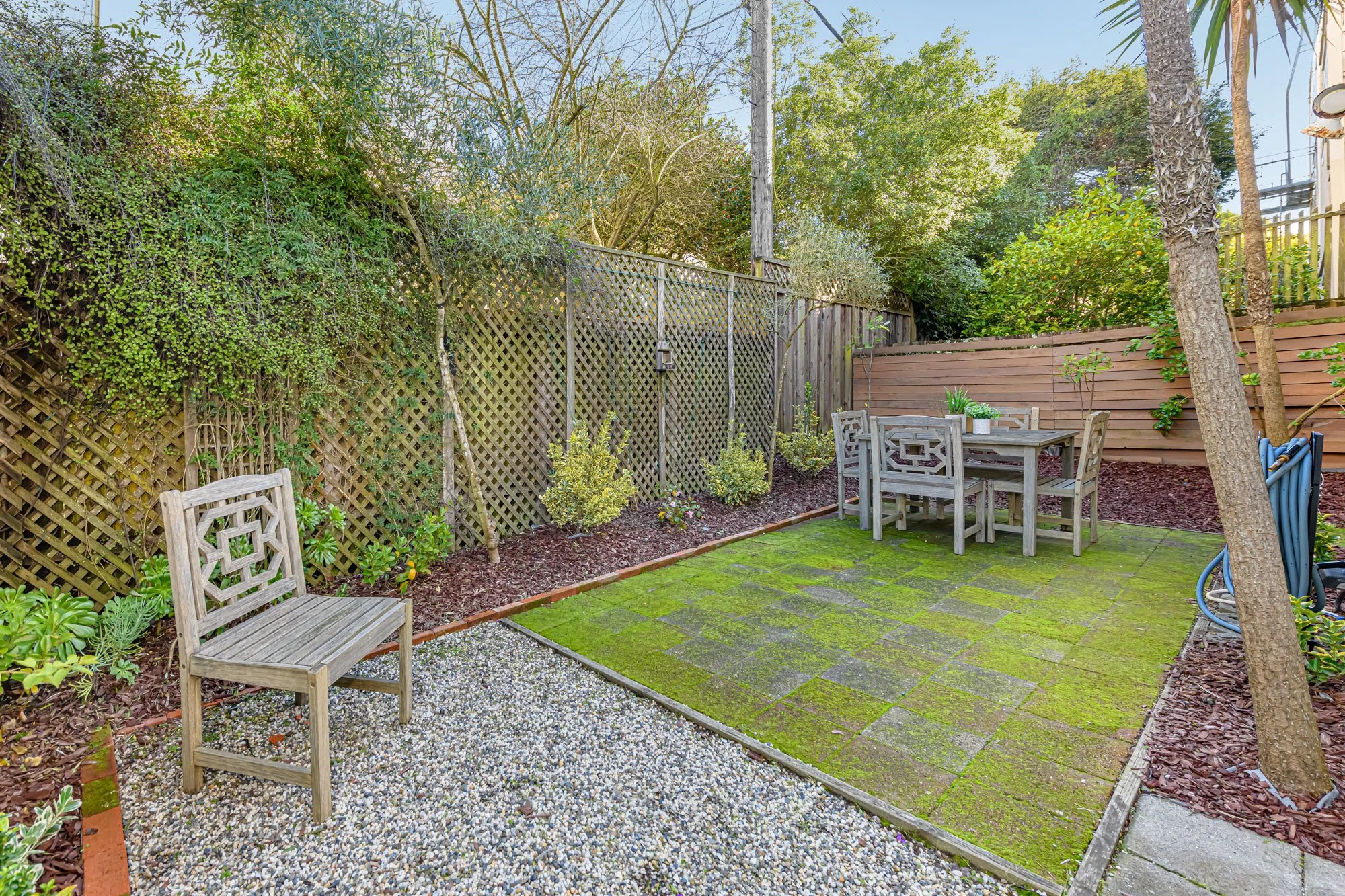 A small outdoor backyard patio with a brick-paved section covered in green moss, bordered by a strip of gravel, and a second section with a wood deck. There are several wooden garden chairs and a table with plants on it. The yard is enclosed with a wooden and lattice fence, with trees and shrubs creating a lush background.