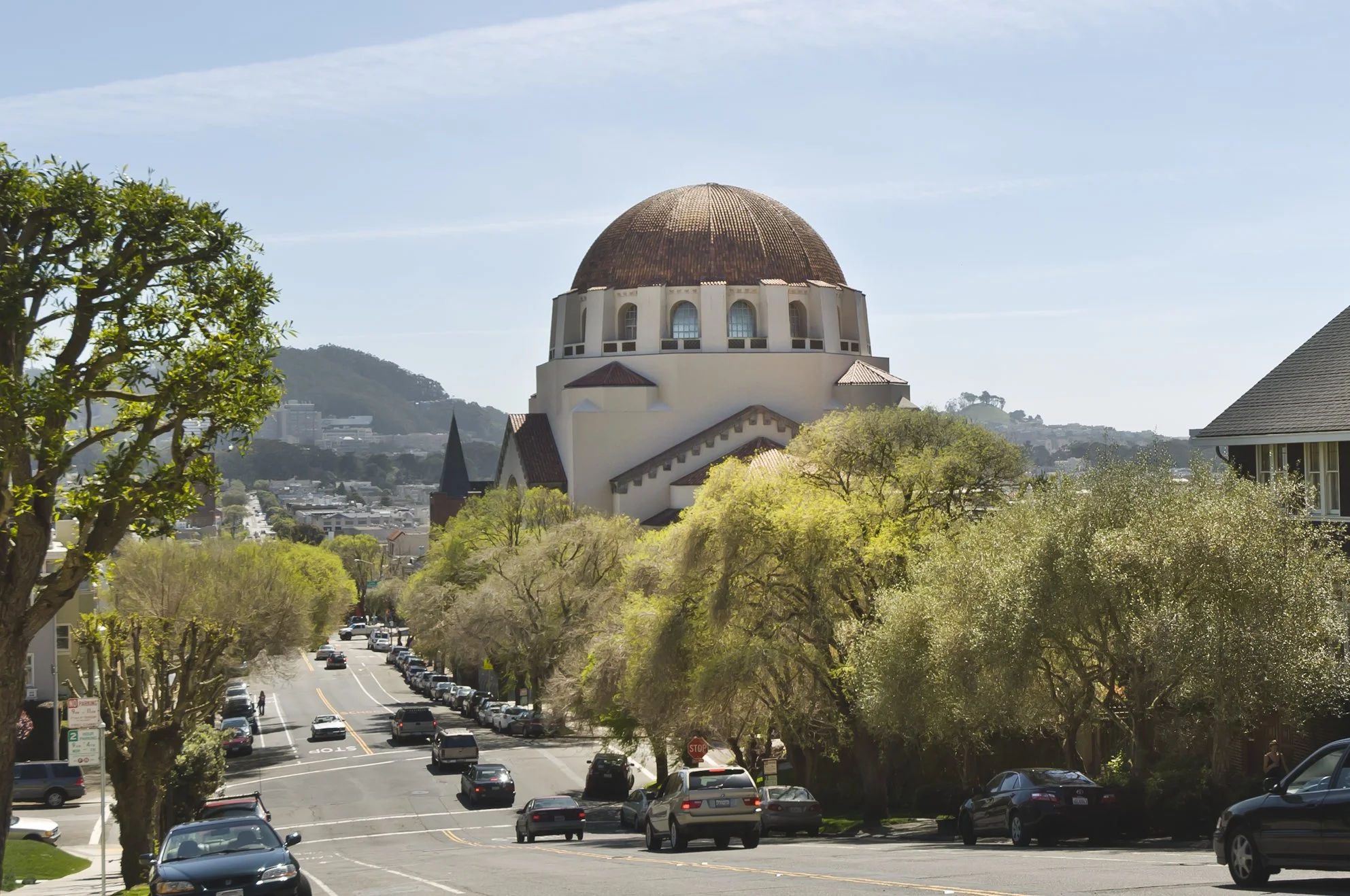 Street view with cars parked along the sides, lined with trees and a large building with a domed roof in the background.
