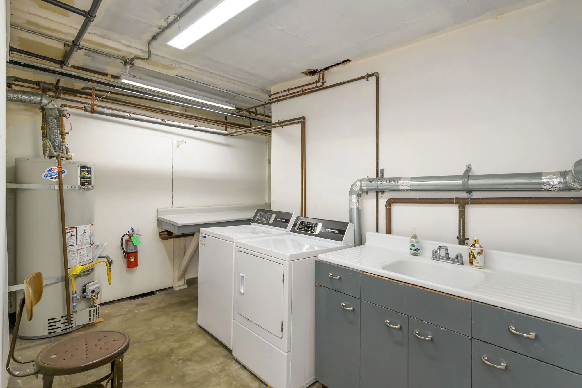 Laundry room with washing machine, utility sink, water heater, fire extinguisher, plastic chair, and visible plumbing and electrical pipes on ceiling and wall.