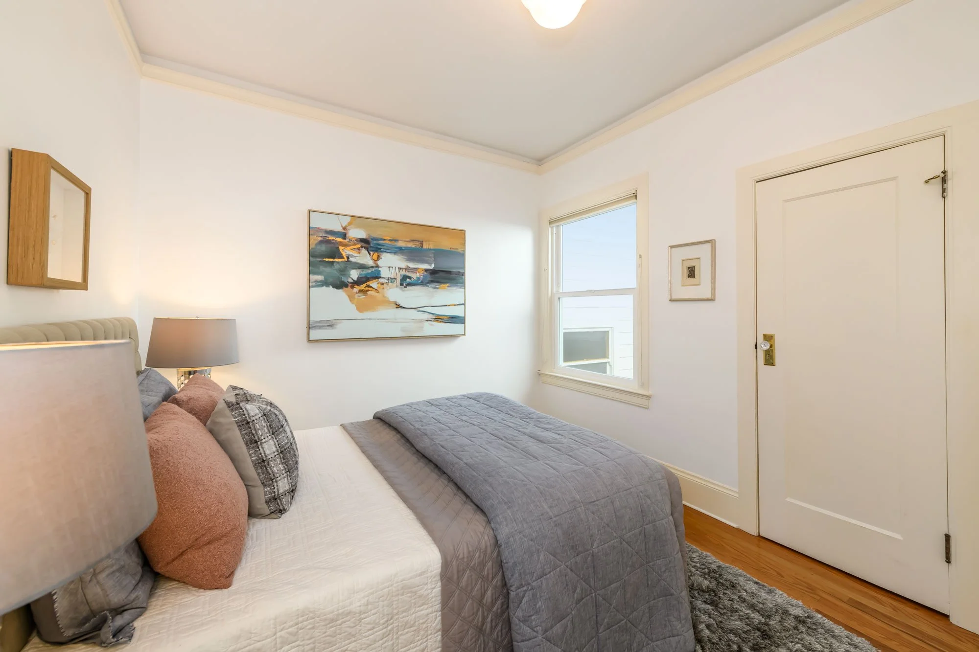 Small bedroom with a bed, several pillows, a gray quilt, and two table lamps. White walls with a painting and window with a view outside. Wooden door and hardwood floor.