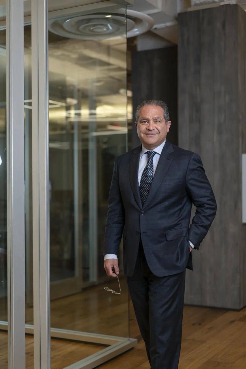 A confident man in a dark suit with a tie, standing in a modern office with glass walls and wood flooring, holding glasses in his right hand and smiling at the camera.