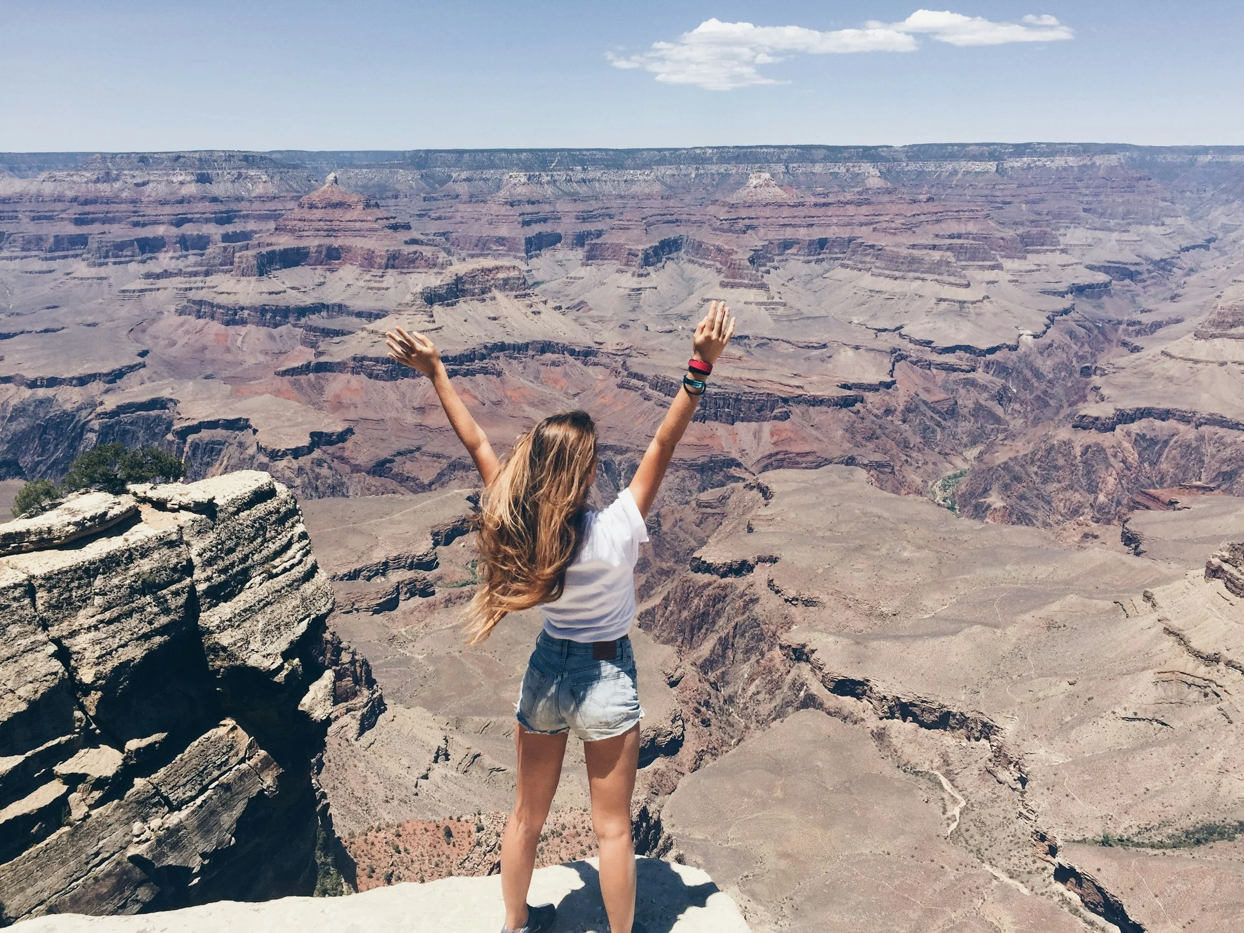 A woman with long hair, wearing a white t-shirt and denim shorts, stands on the edge of the Grand Canyon with arms raised in the air.
