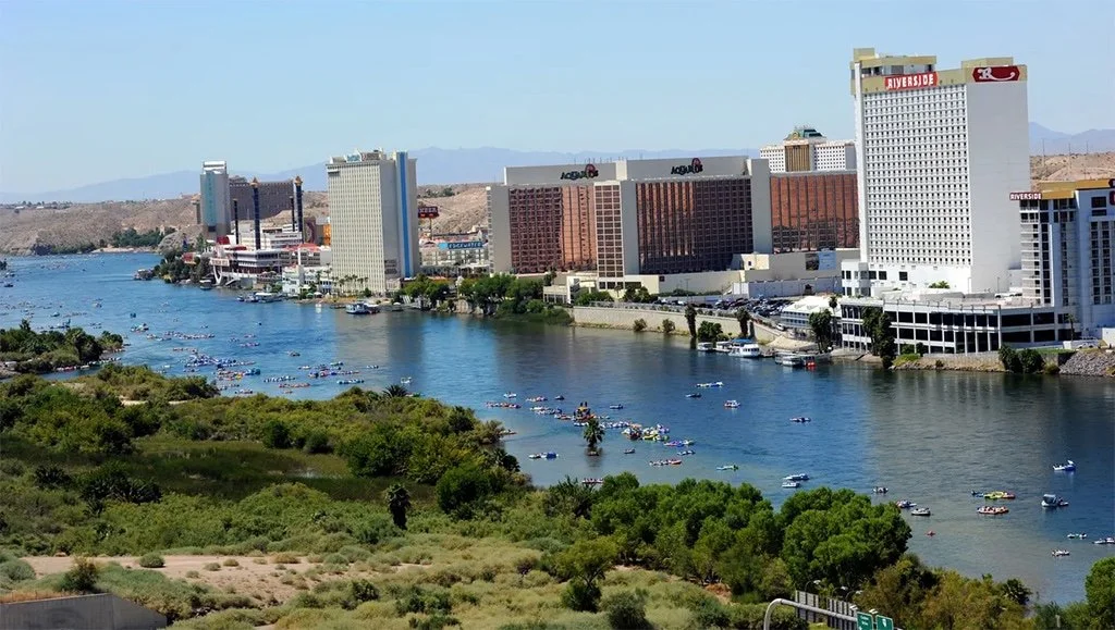 View of a city skyline with high-rise hotels and casinos along a river, with boats on the water and green vegetation in the foreground, likely in Las Vegas.