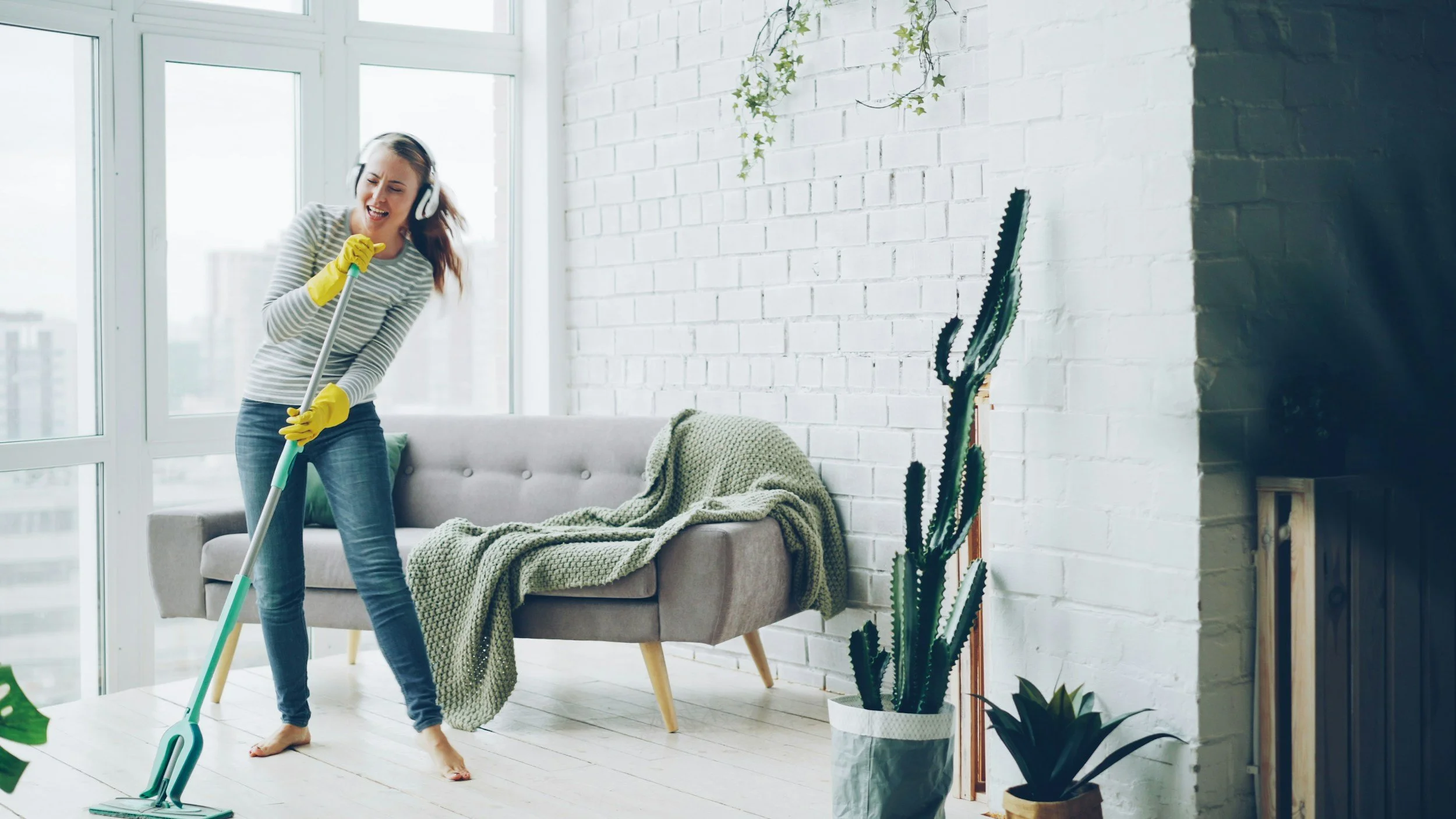 Woman cleaning the floor with a mop in a bright, modern living room with large windows and potted plants.