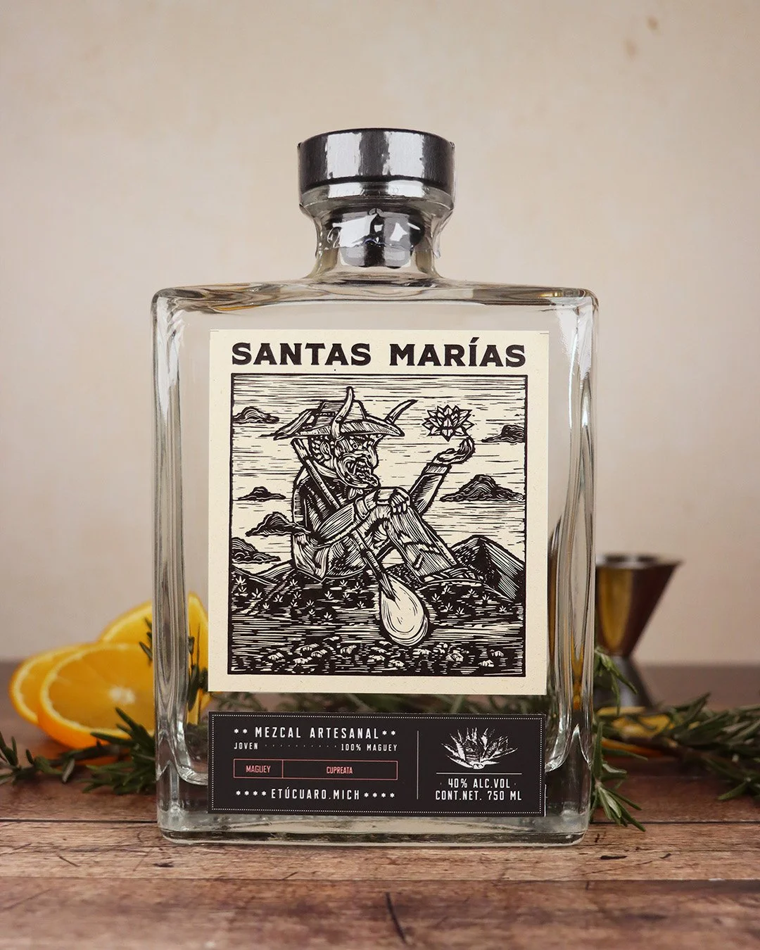 Botella de mezcal artesanal llamada 'Santas Marías' con etiqueta en negro y blanco, ilustración de un diablillo con cuernos, sentado en una roca, sosteniendo una flor. Rodeada de limones, romero y un molinillo como fondo, en una mesa de madera.