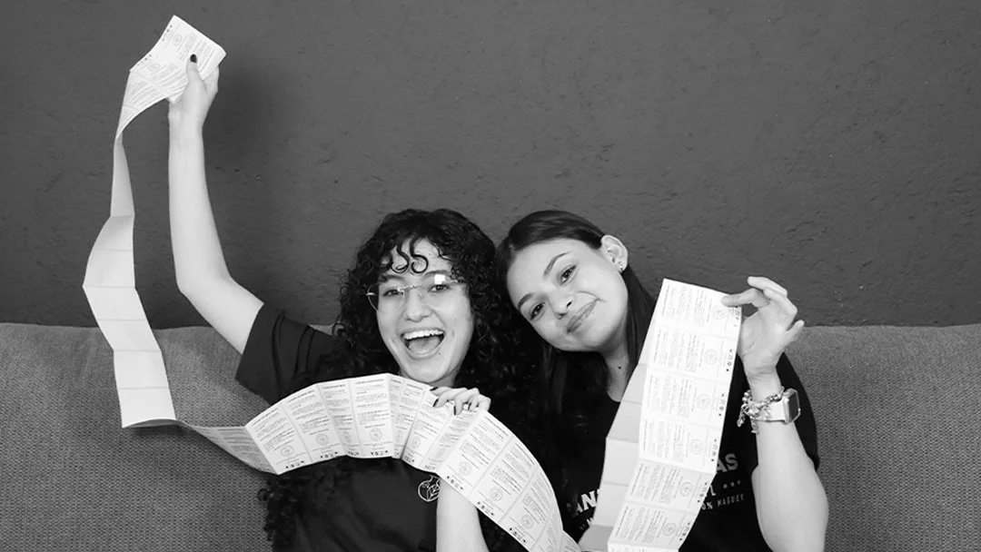 Dos mujeres sonrientes sosteniendo y mostrando boletos o tarjetas largas y plegadas, sentadas en un sillón contra una pared de fondo.