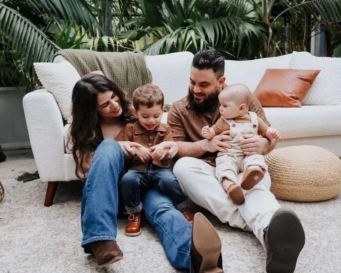 Family of four sitting on the floor relaxing in front of a white sofa with green plants in the background. Mother, father, and two young children laughing and playing together.