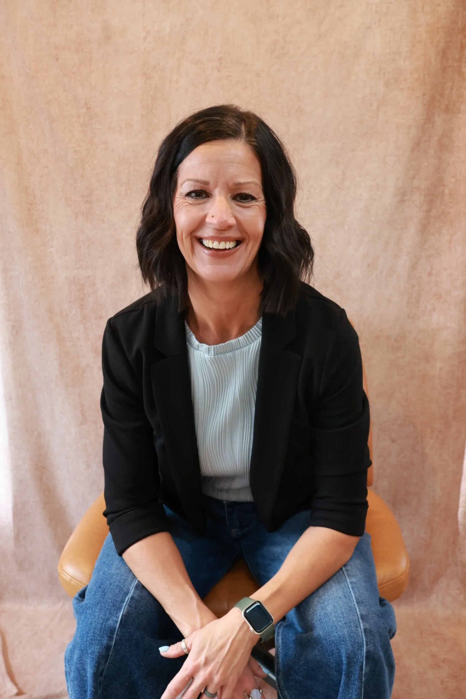 A woman with shoulder-length black hair, wearing a black blazer, white ribbed top, blue jeans, and a smartwatch, sitting on a tan chair and smiling at the camera, against a beige background.